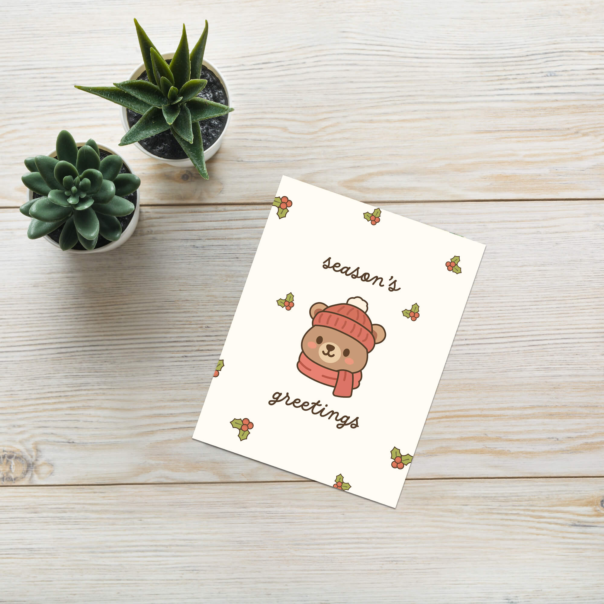 cute Christmas card with a bear wearing a pink hat and scarf, surrounded by holly leaves and berries with text: season's greetings
