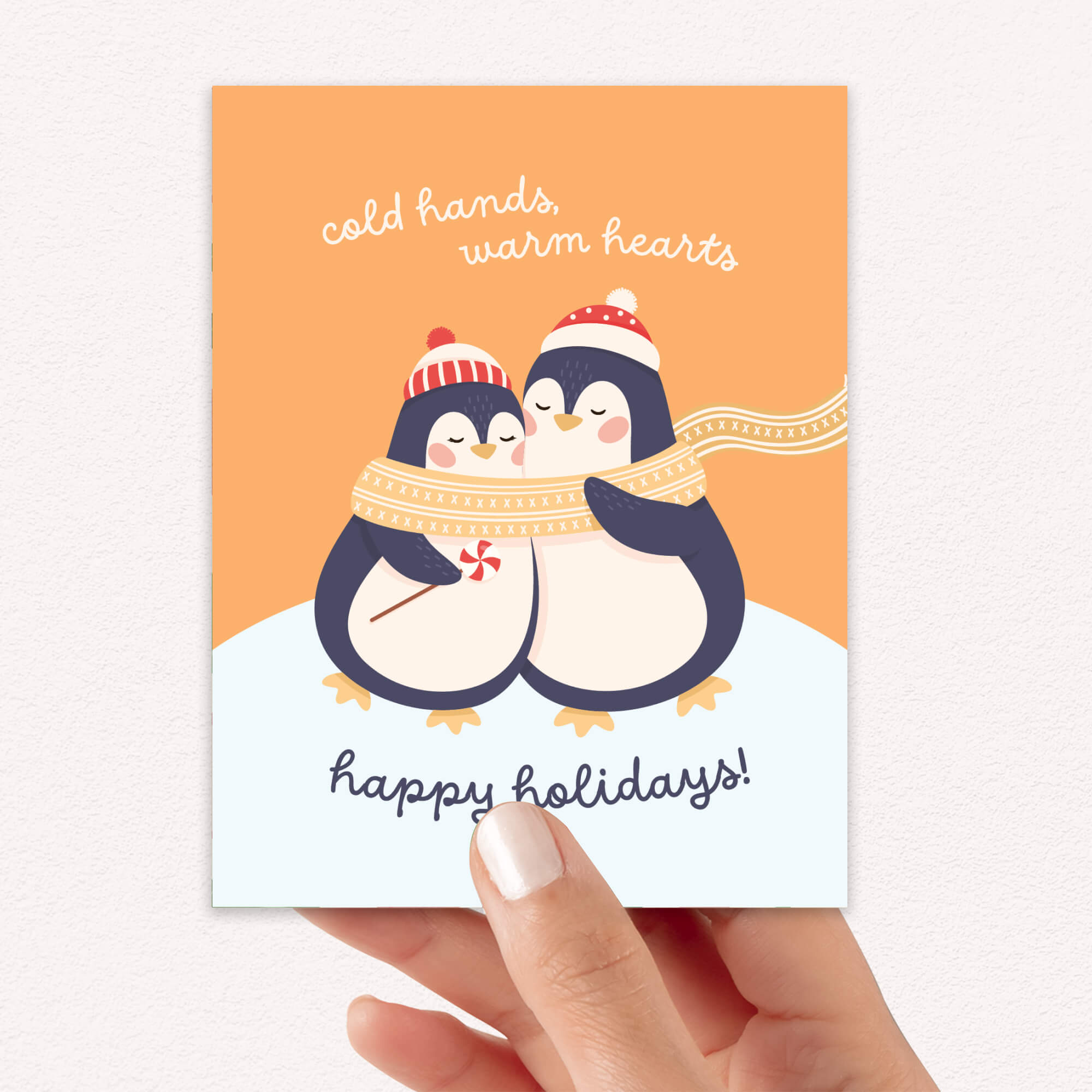 unique holiday card with two penguins hugging, wearing scarves and hats, on an orange background with text "Two penguins in knit hats sharing a scarf; text “cold hands, warm hearts — happy holidays!”"