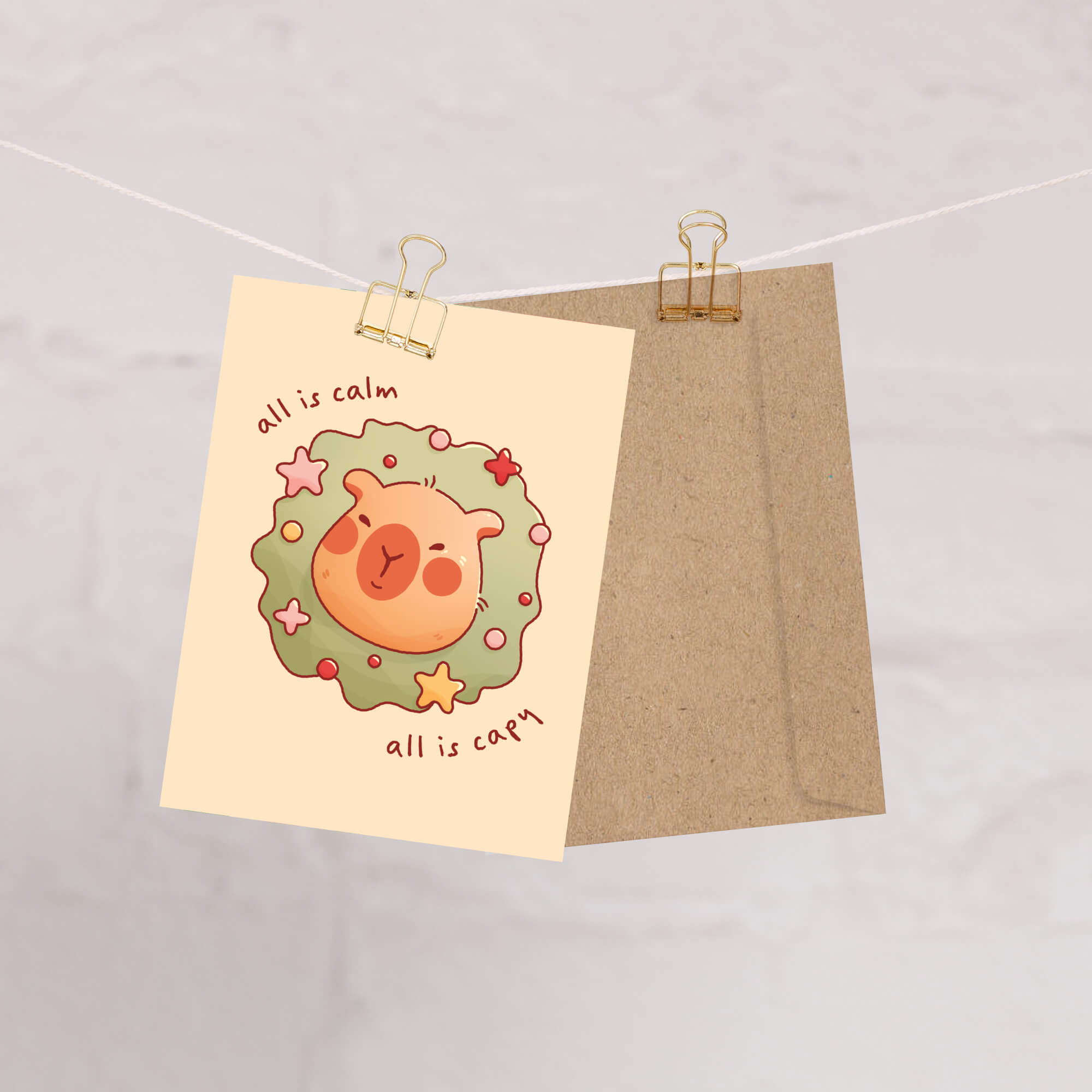 cute christmas card with a Capybara nestled in green holiday wreath with stars, text “all is calm, all is capy.”