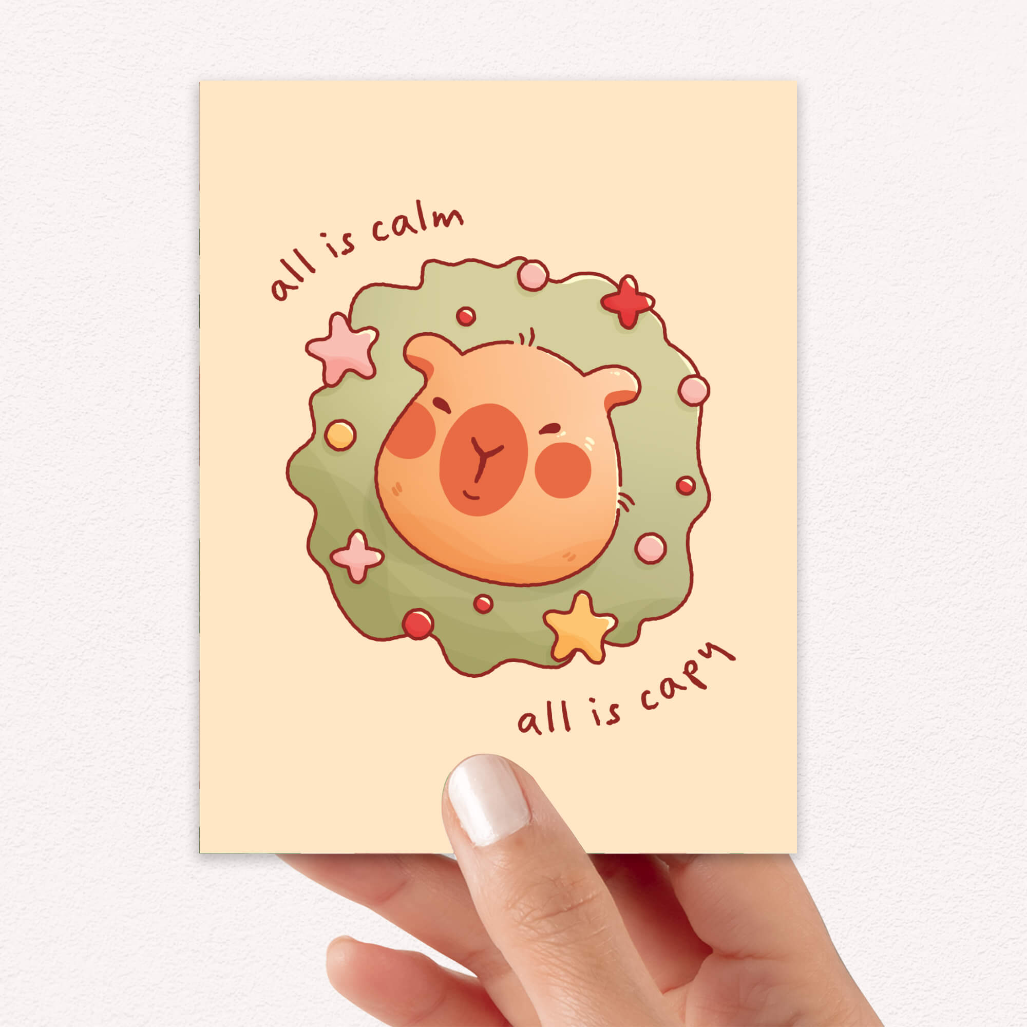 cute christmas card with a Capybara nestled in green holiday wreath with stars, text “all is calm, all is capy.”