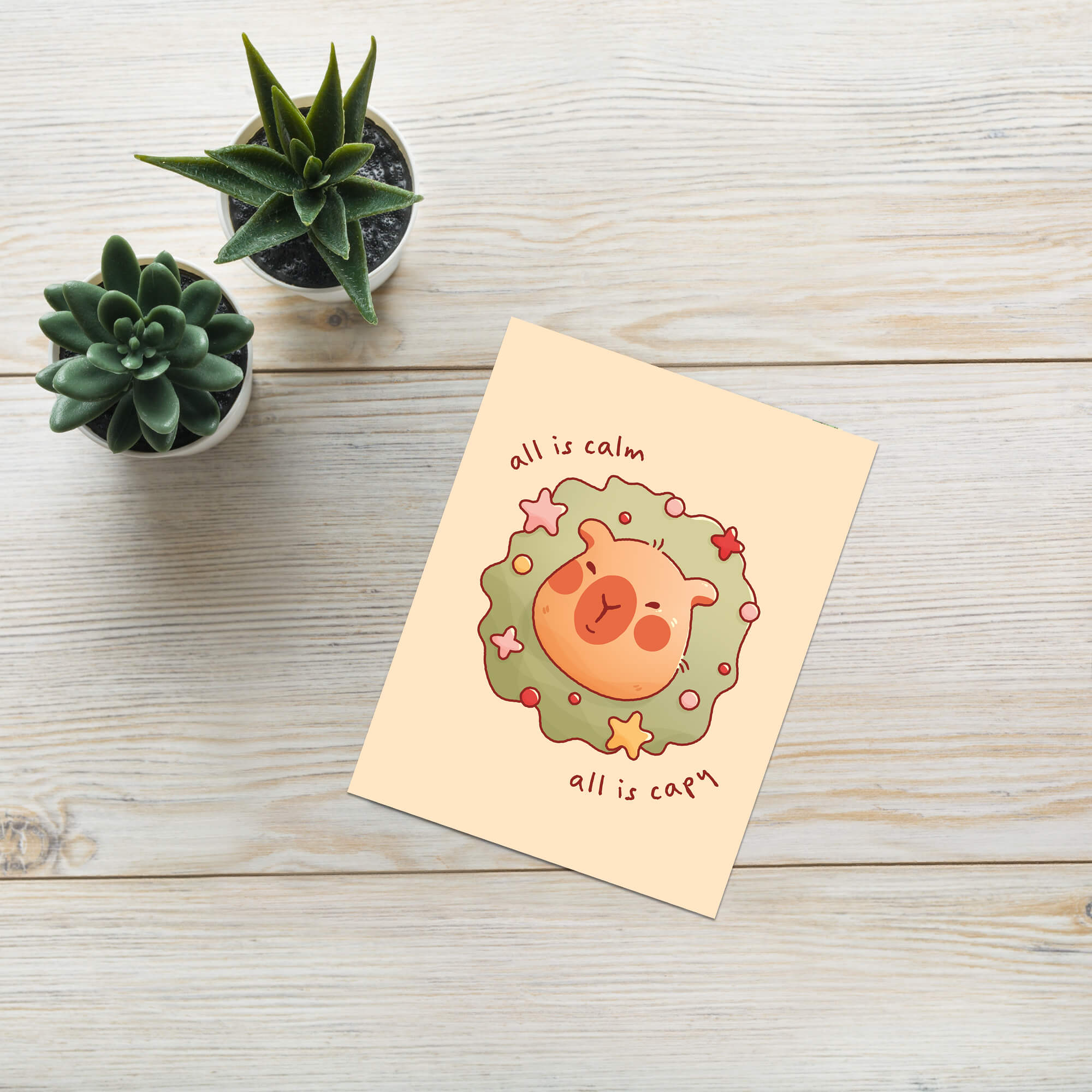 cute christmas card with a Capybara nestled in green holiday wreath with stars, text “all is calm, all is capy.”
