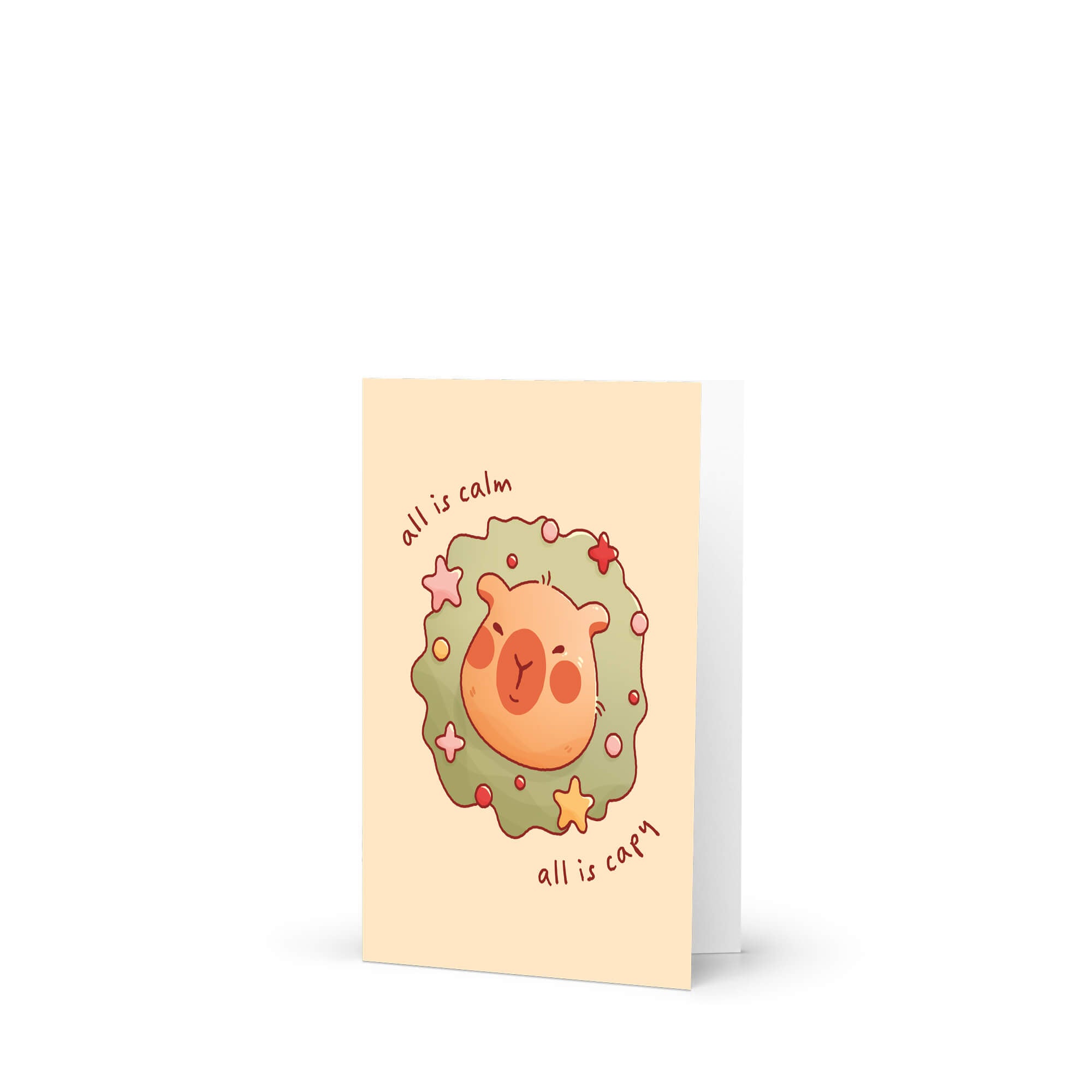 cute christmas card with a Capybara nestled in green holiday wreath with stars, text “all is calm, all is capy.”