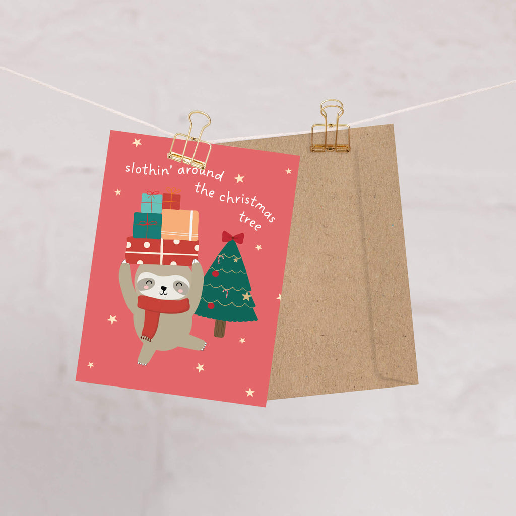 unique Christmas card with a sloth carrying presents and a holiday tree on a red background with text "slothin' around the christmas tree"