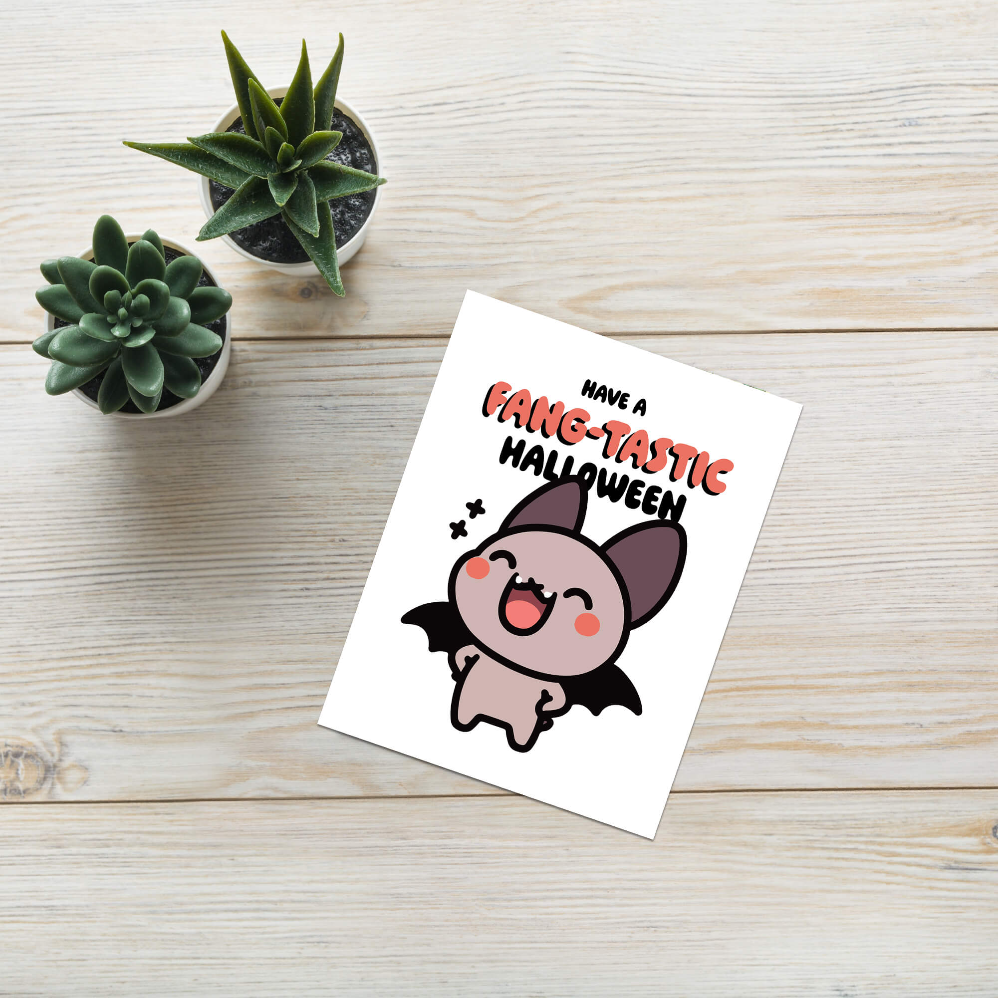 Greeting card with a bat illustration and 'Have a Fang-tastic Halloween' text on a wooden surface with small plants.