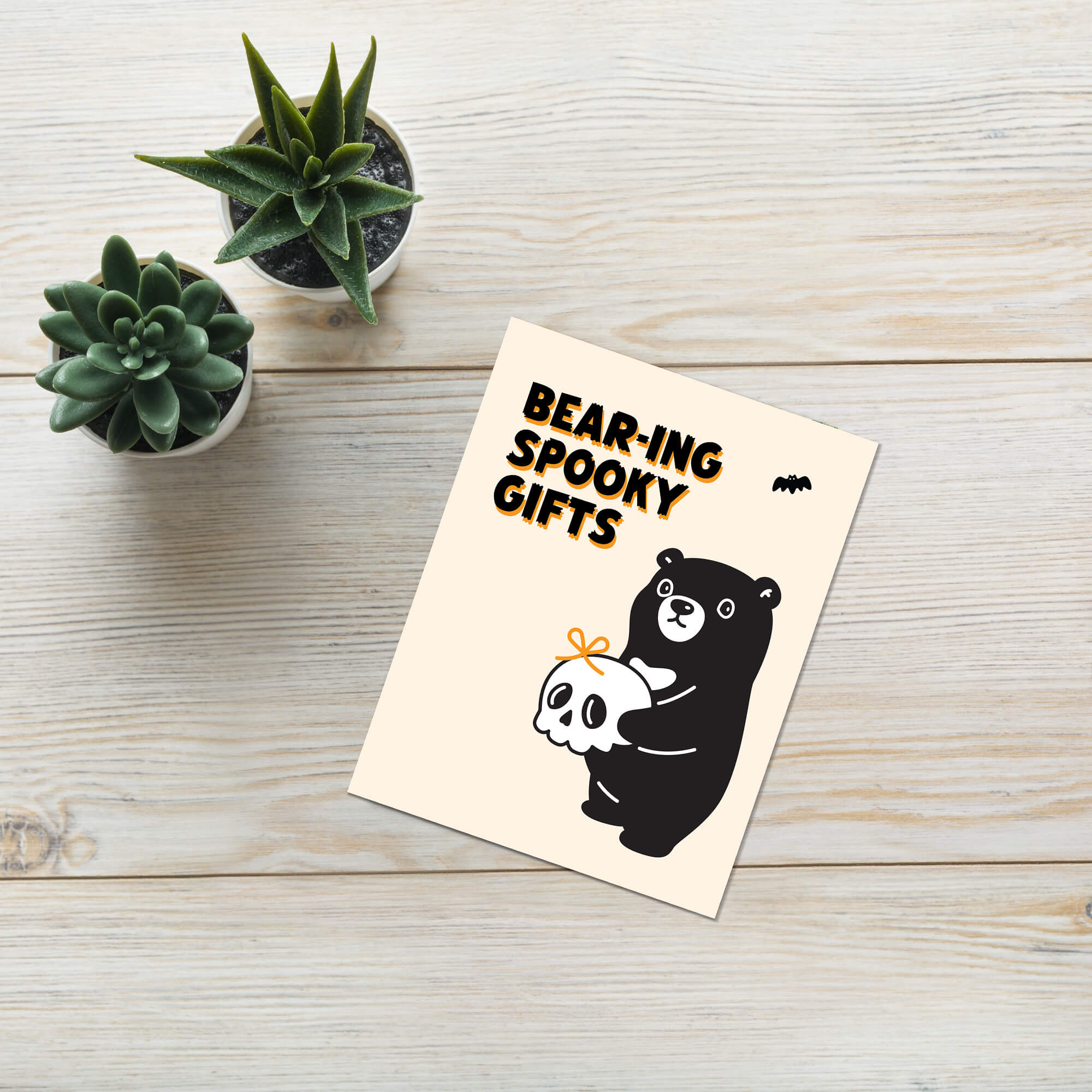 Greeting card with bear and ghost illustration on a wooden surface with plants