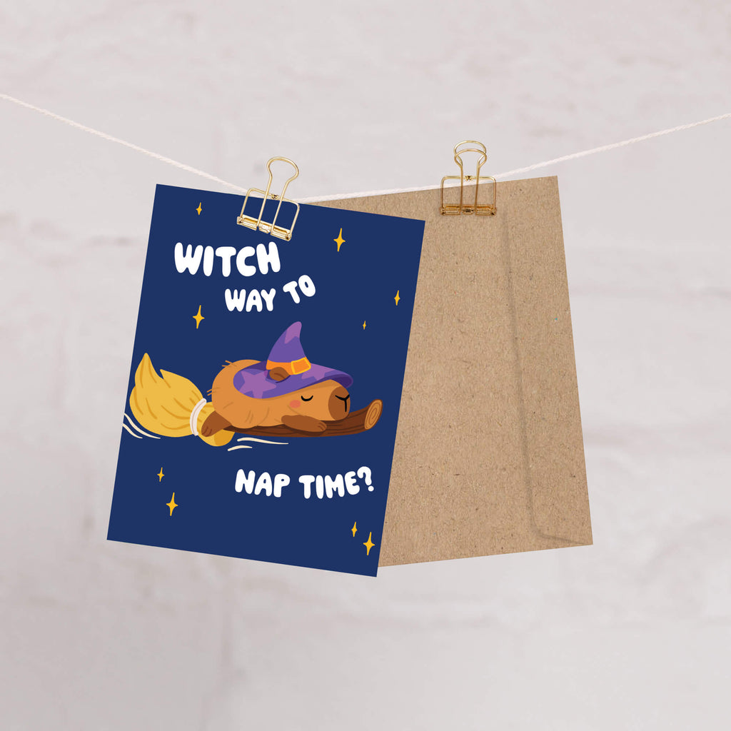 Halloween-themed greeting card with a capybara in a witch hat and broom, text 'Witch way to nap time?' on a blue background, clipped to a string.