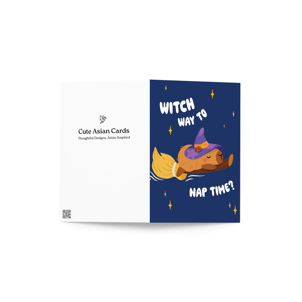 Halloween-themed greeting card with a capybara in a witch hat and broom, text 'Witch way to nap time?' on a blue background, clipped to a string.