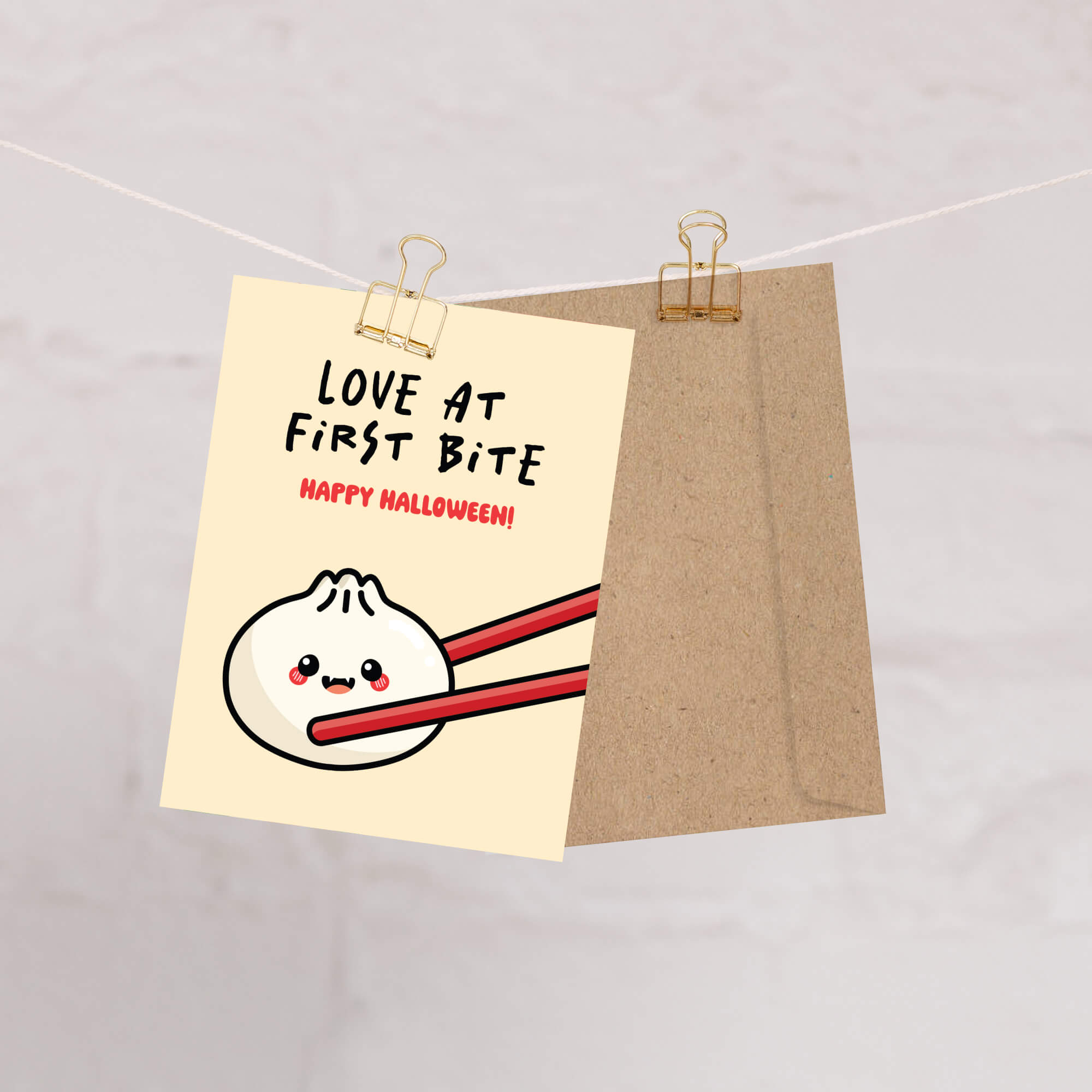 Greeting card with a cartoon dumpling with fangsholding chopsticks and text 'Love at First Bite Happy Halloween!' on a beige background.