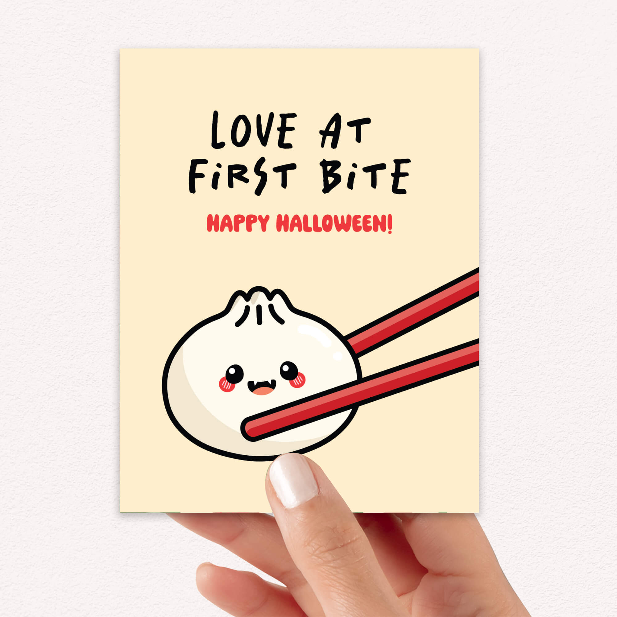 Greeting card with a cartoon dumpling with fangsholding chopsticks and text 'Love at First Bite Happy Halloween!' on a beige background.
