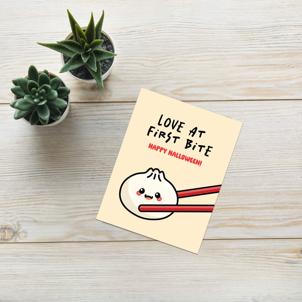 Greeting card with a cartoon dumpling with fangsholding chopsticks and text 'Love at First Bite Happy Halloween!' on a beige background.
