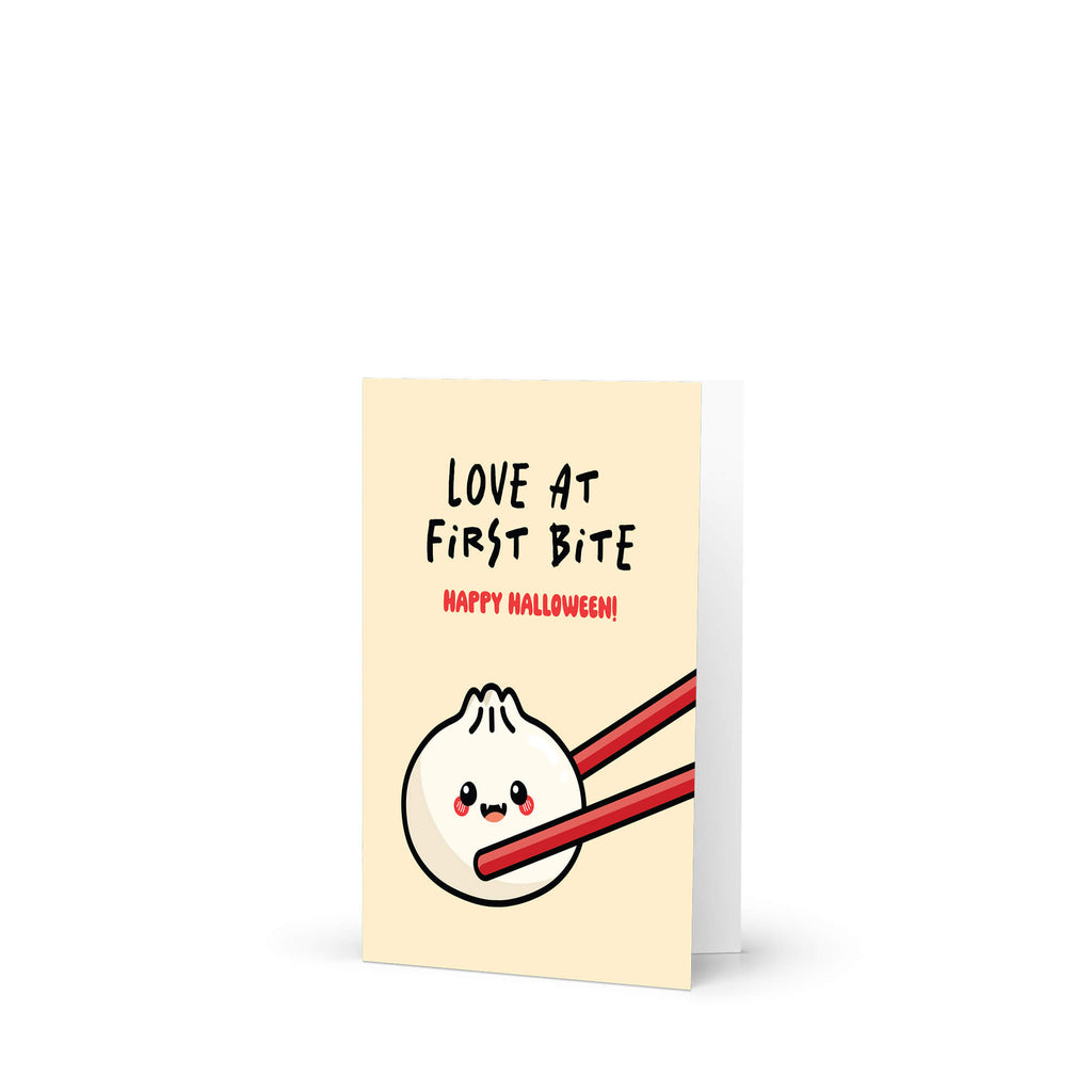 Greeting card with a cartoon dumpling with fangsholding chopsticks and text 'Love at First Bite Happy Halloween!' on a beige background.