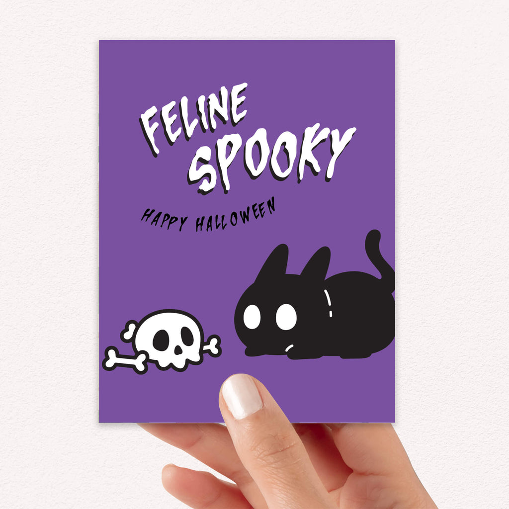 Halloween card with black cat and skull on a purple background held by a hand.