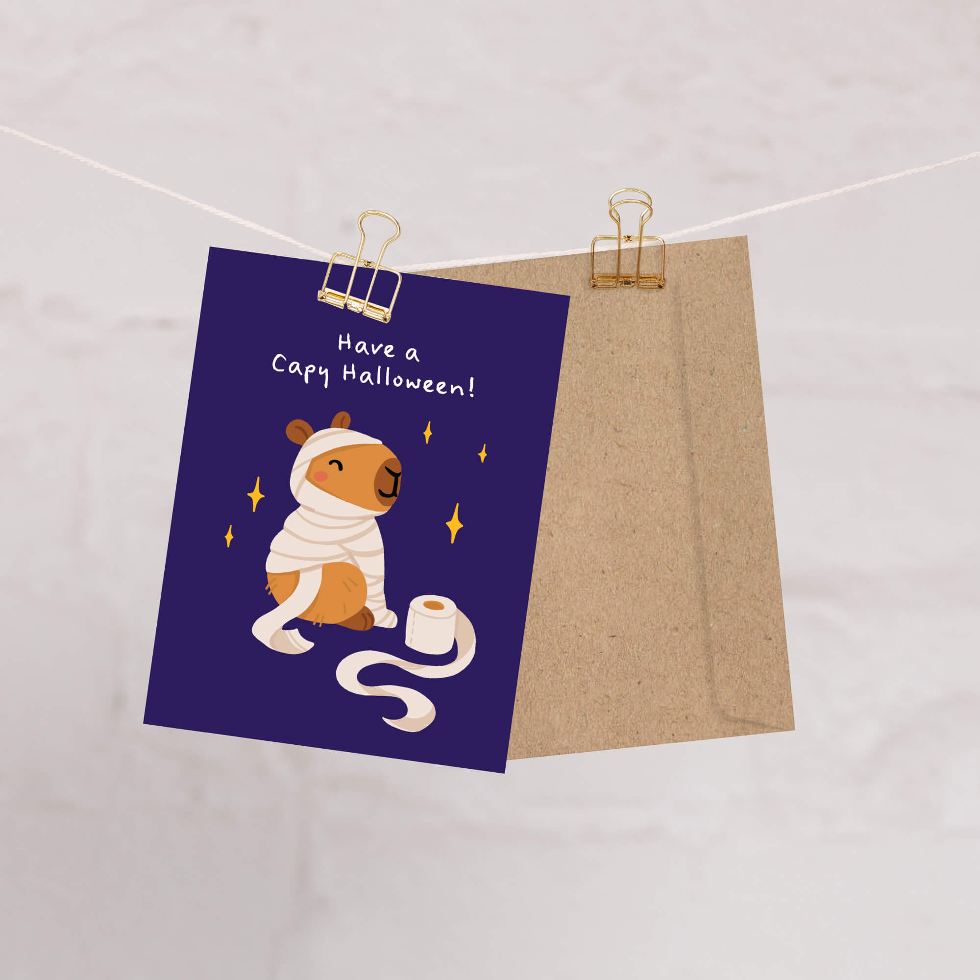 Halloween card with a capybara illustration and text on a purple background, clipped to a string.