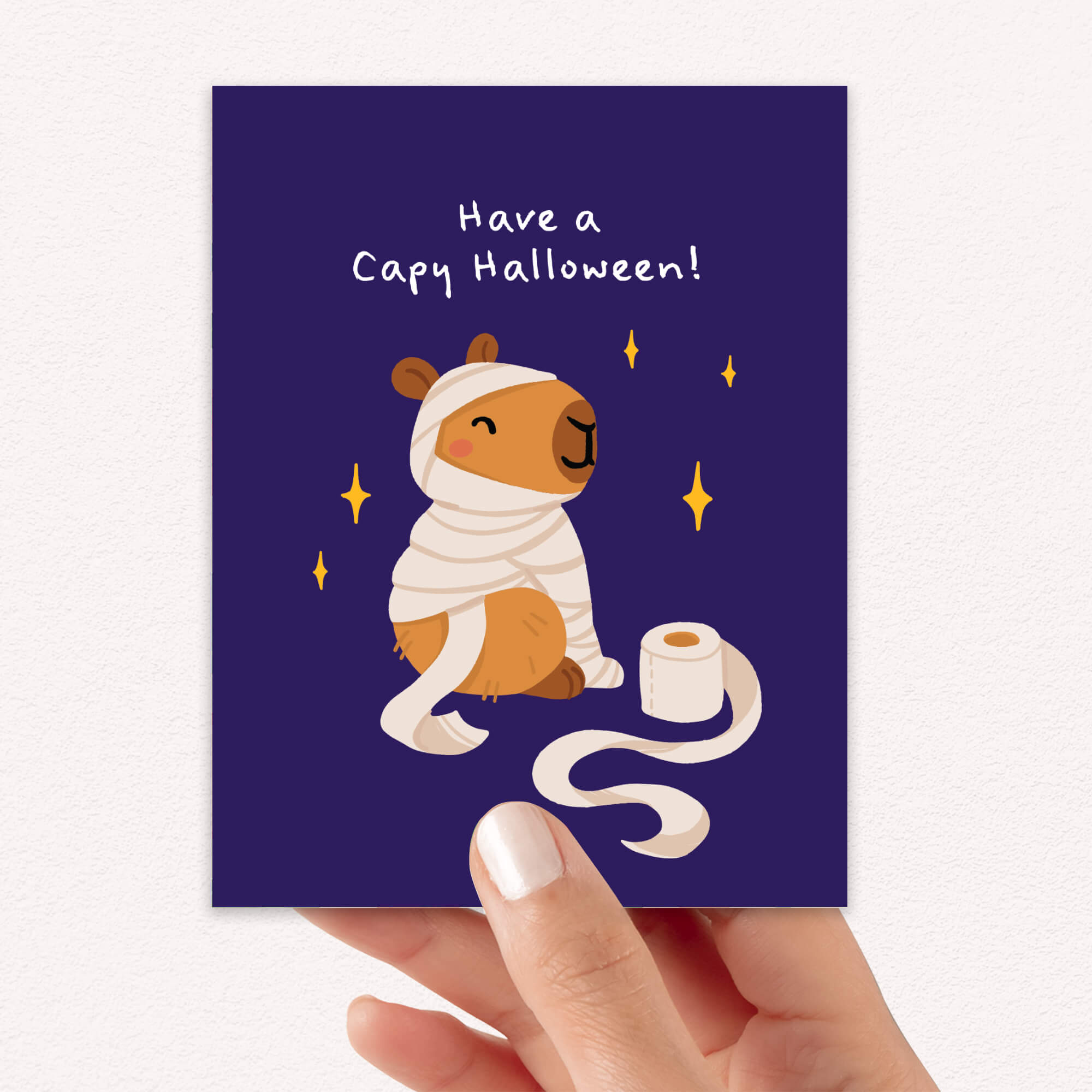 Halloween card with a capybara wearing a cape and holding toilet paper on a dark blue background.