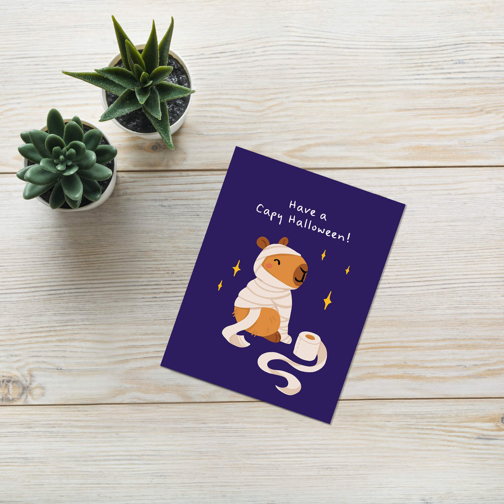 Greeting card with a bear costume illustration and 'Have a Capy Halloween!' text on a wooden surface with plants.