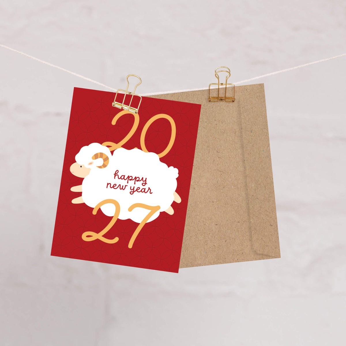 A lunar new year card with gold '2027' numbers and a white sheep graphic reading 'happy new year,' clipped to a string next to a kraft envelope on a light background.