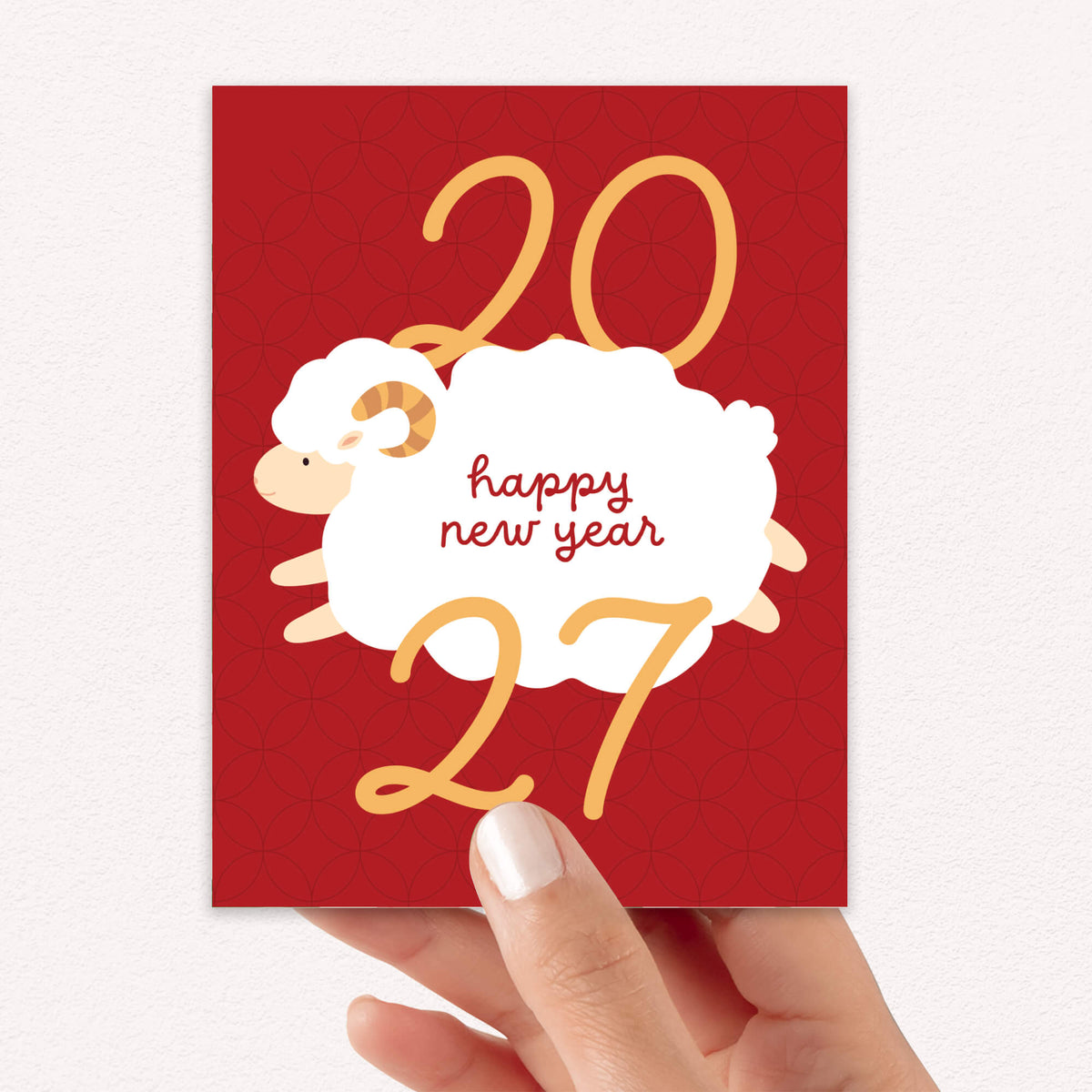 A hand holding a year of the sheep card featuring a white ram illustration, gold numbers '20' and '27', and the words 'happy new year' in red script.