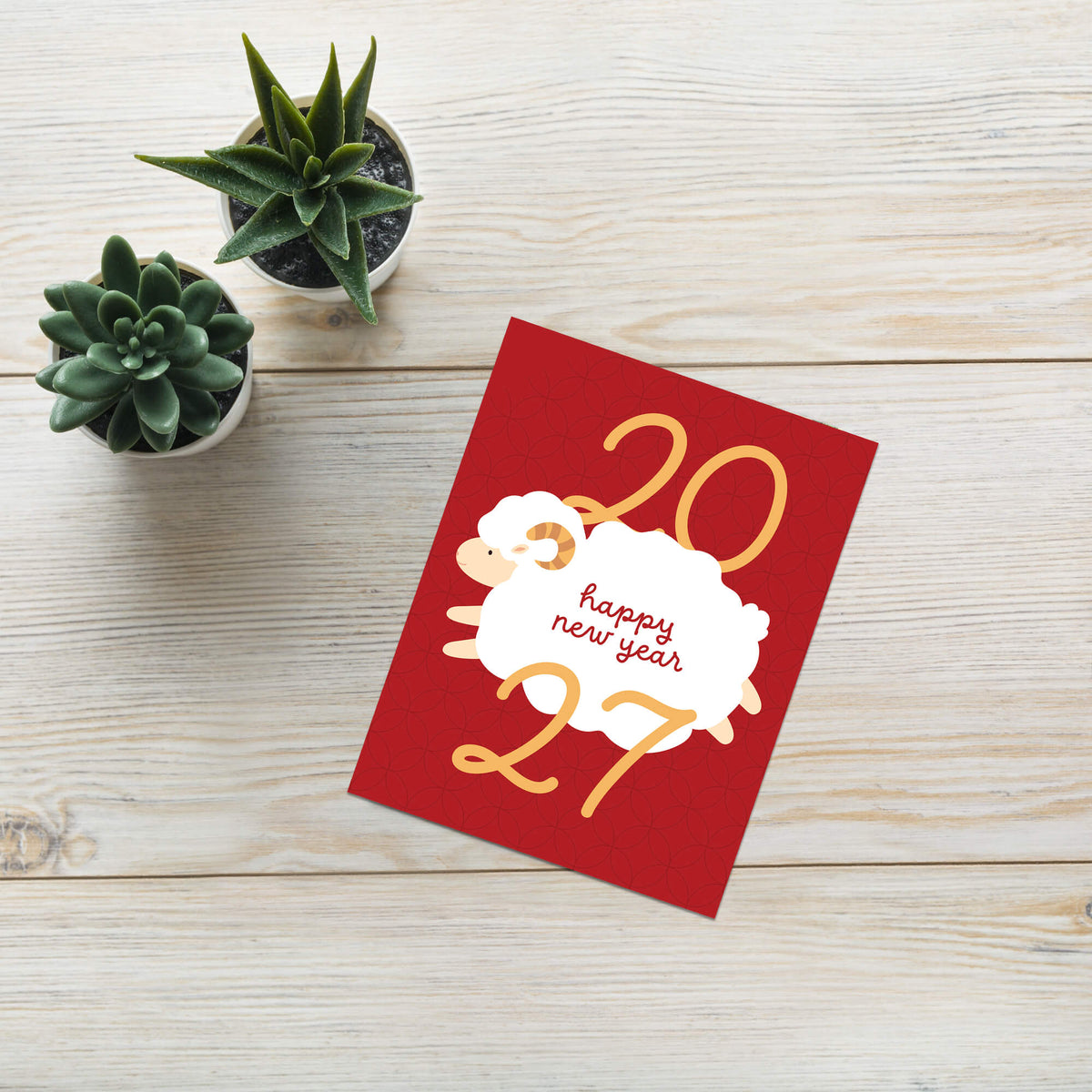 A red year of the ram card with a white sheep illustration and gold numbers reading '2027' and 'happy new year', placed on a light wood surface next to two small potted succulents.