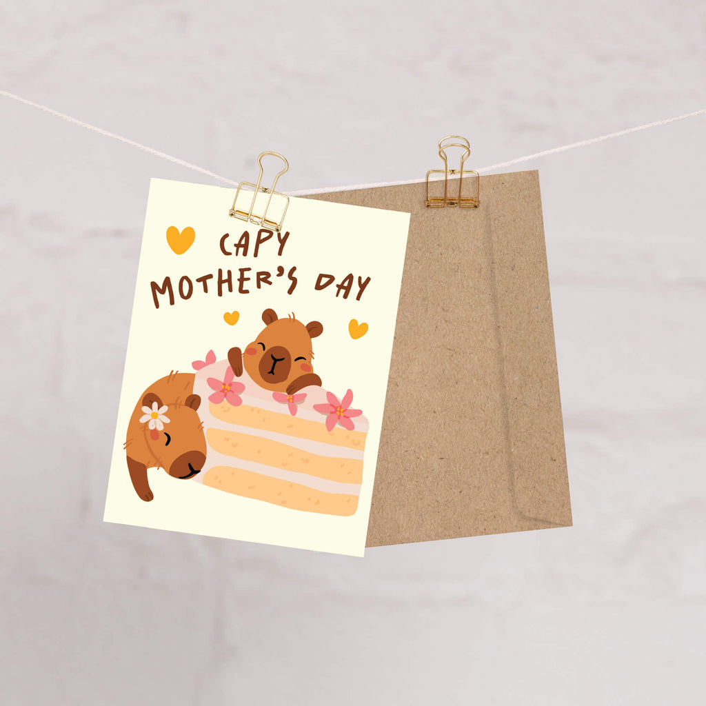 Mother's Day card with cute capybara animals around a cake light on a light yellow background with funny pun message: capy mother's day surrounded by hearts