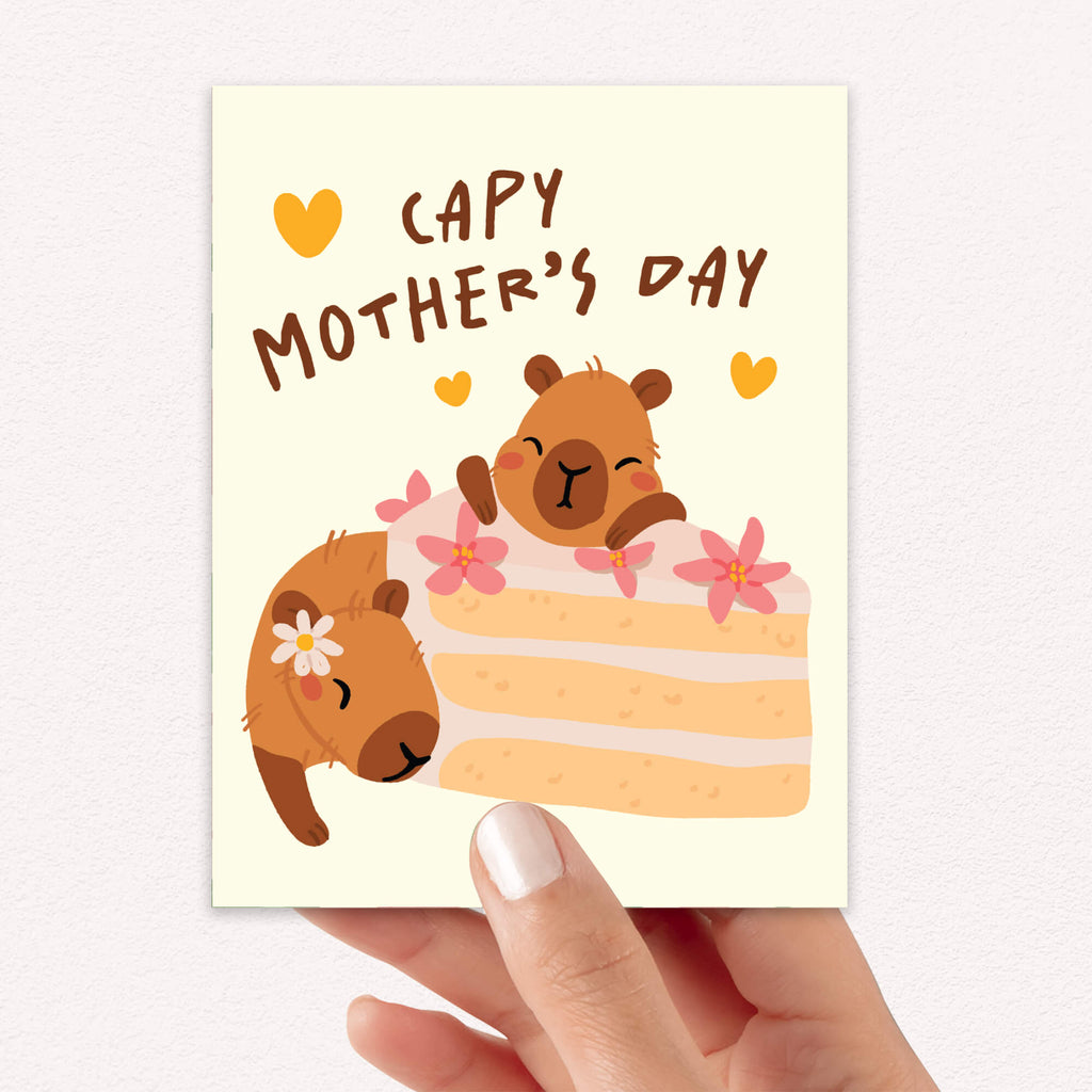 Mother's Day card with cute capybara animals around a cake light on a light yellow background with funny pun message: capy mother's day surrounded by hearts
