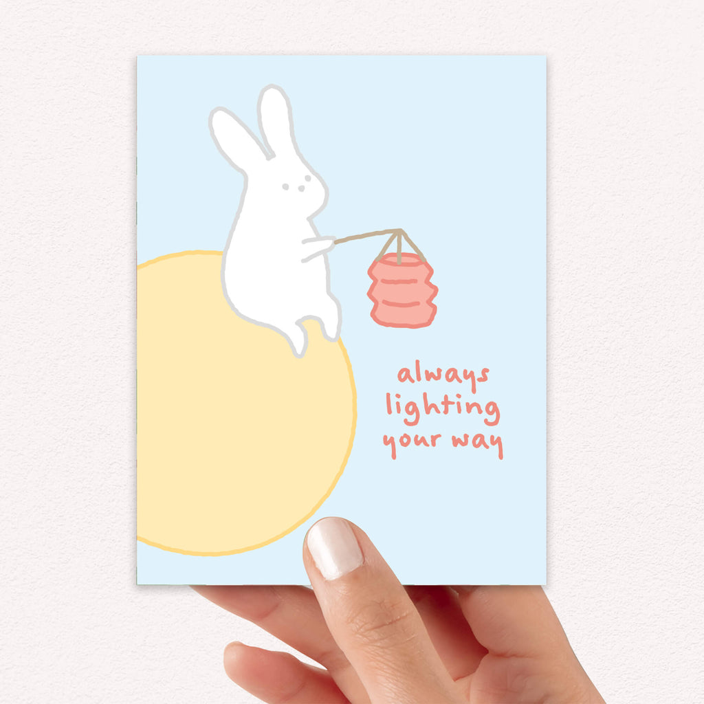 asian inspired Greeting card with a rabbit holding a lantern and text 'always lighting your way' on a light blue background. for mid autumn festival