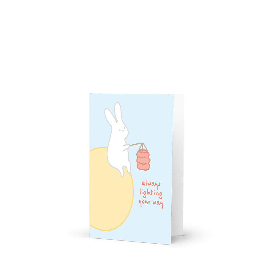 asian inspired Greeting card with a rabbit holding a lantern and text 'always lighting your way' on a light blue background. for mid autumn festival