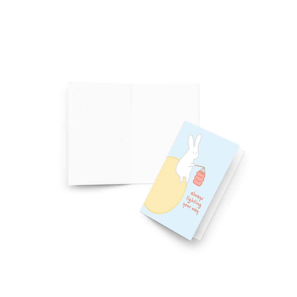 asian inspired Greeting card with a rabbit holding a lantern and text 'always lighting your way' on a light blue background. for mid autumn festival