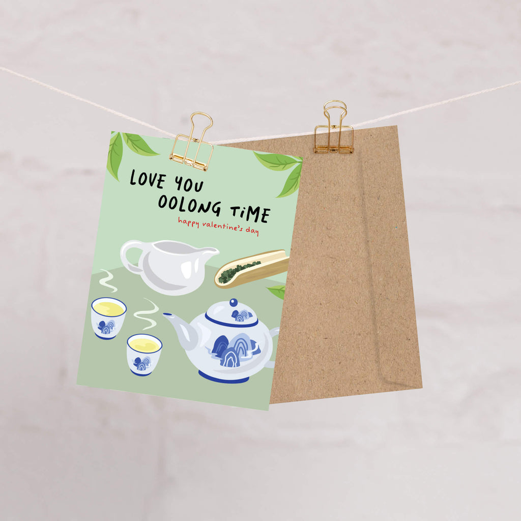 asian inspired valentintues day card with White and blue china teapot, cups, and oolong loose leaf tea on soft green; text “Love you oolong time.”