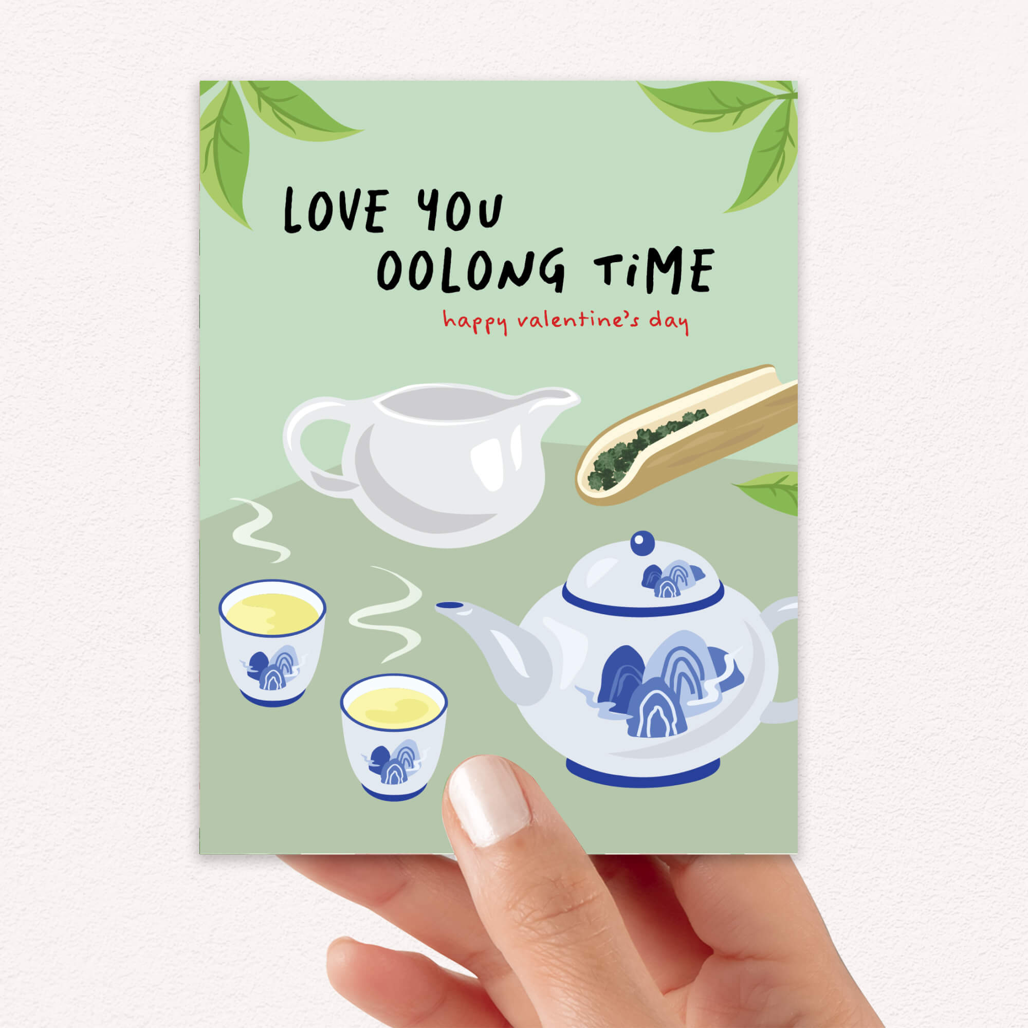 asian inspired valentintues day card with White and blue china teapot, cups, and oolong loose leaf tea on soft green; text “Love you oolong time.”