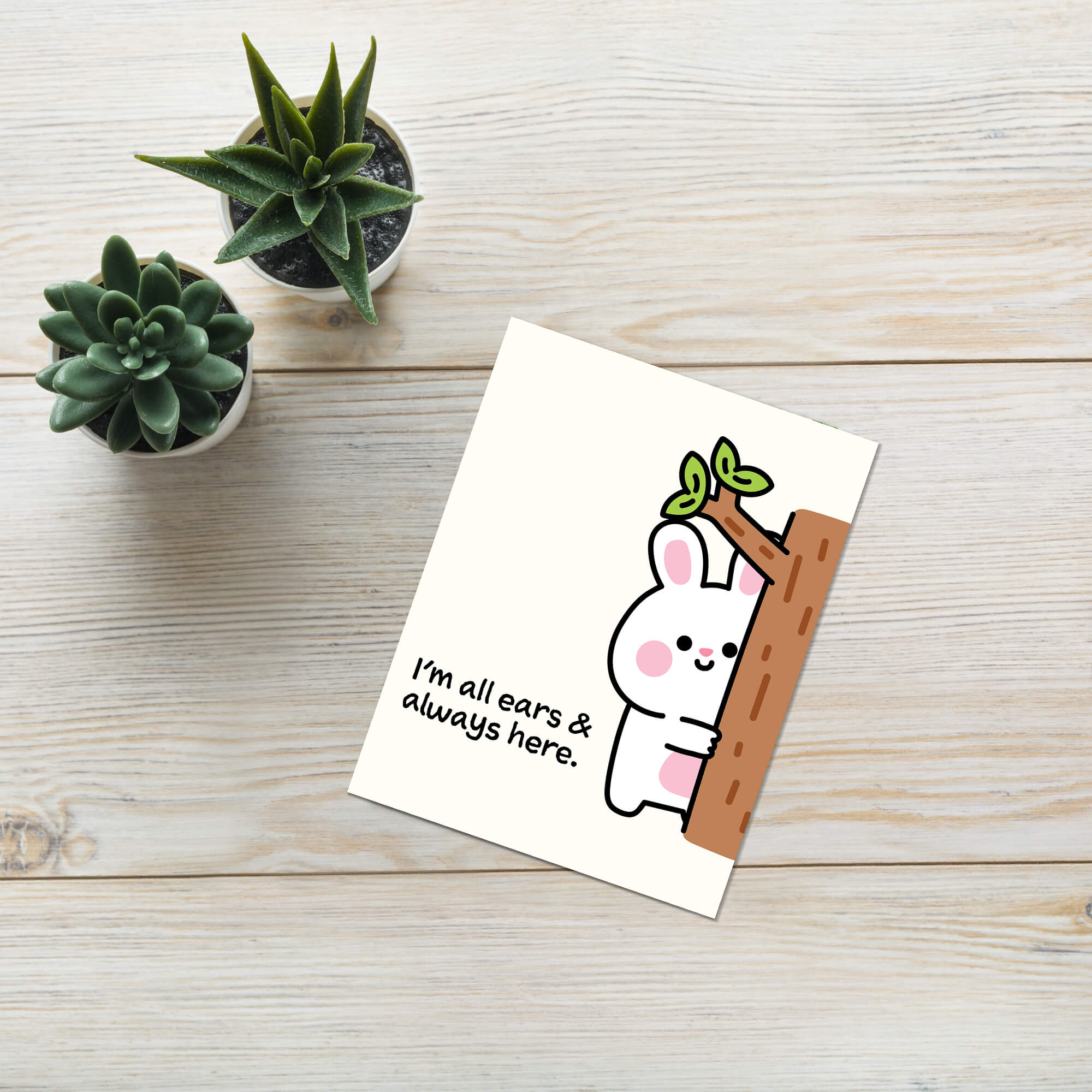 Card with a cartoon rabbit holding a branch on a wooden surface with small plants.