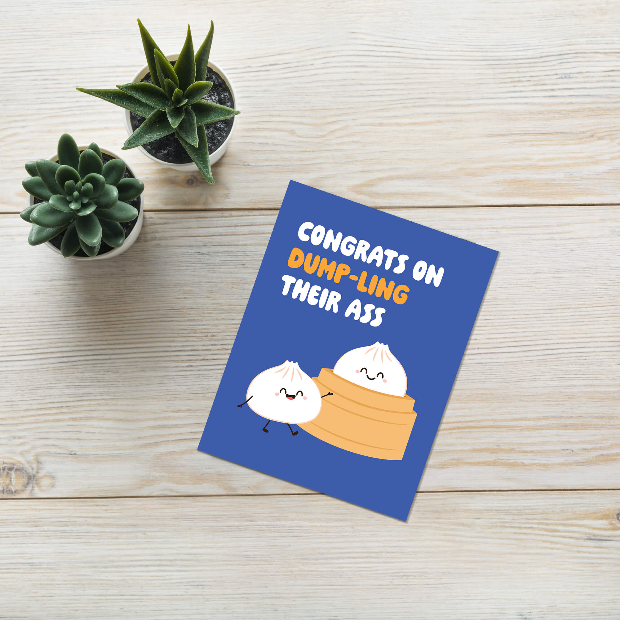 Greeting card with cartoon dumplings and humorous text on a wooden surface with plants.