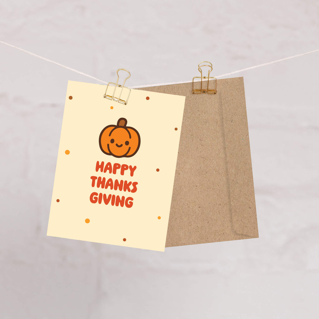 Card with a pumpkin and 'Happy Thanksgiving' text on beige paper, attached to a brown envelope with gold clips.