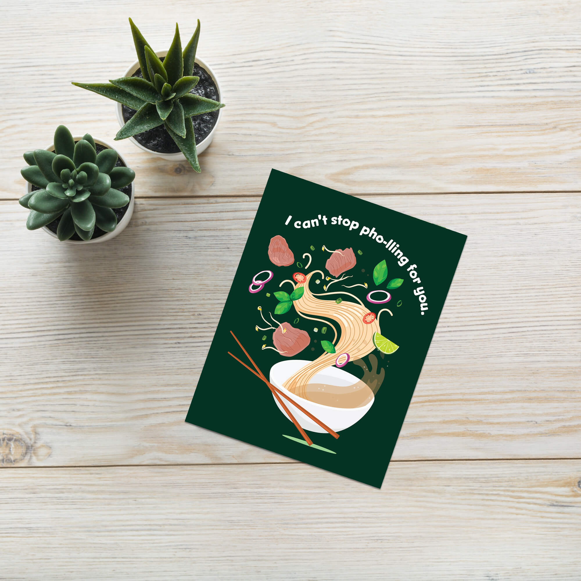 Greeting card with illustration of noodles and text 'I can't stop pho-ling for you.' on a wooden surface with potted plants.