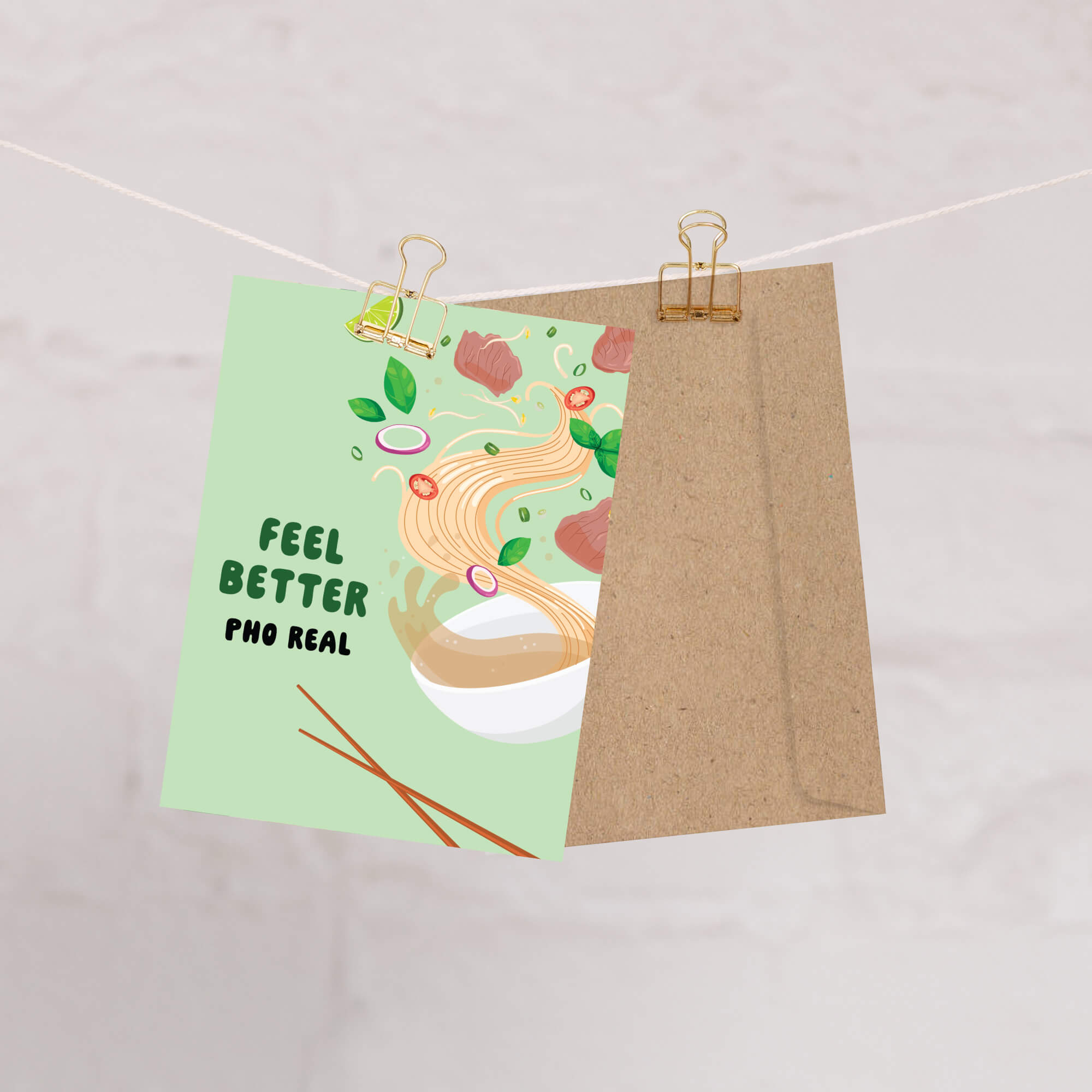 Card with 'Feel Better Pho Real' text and a bowl of pho illustration on a string with a brown envelope.