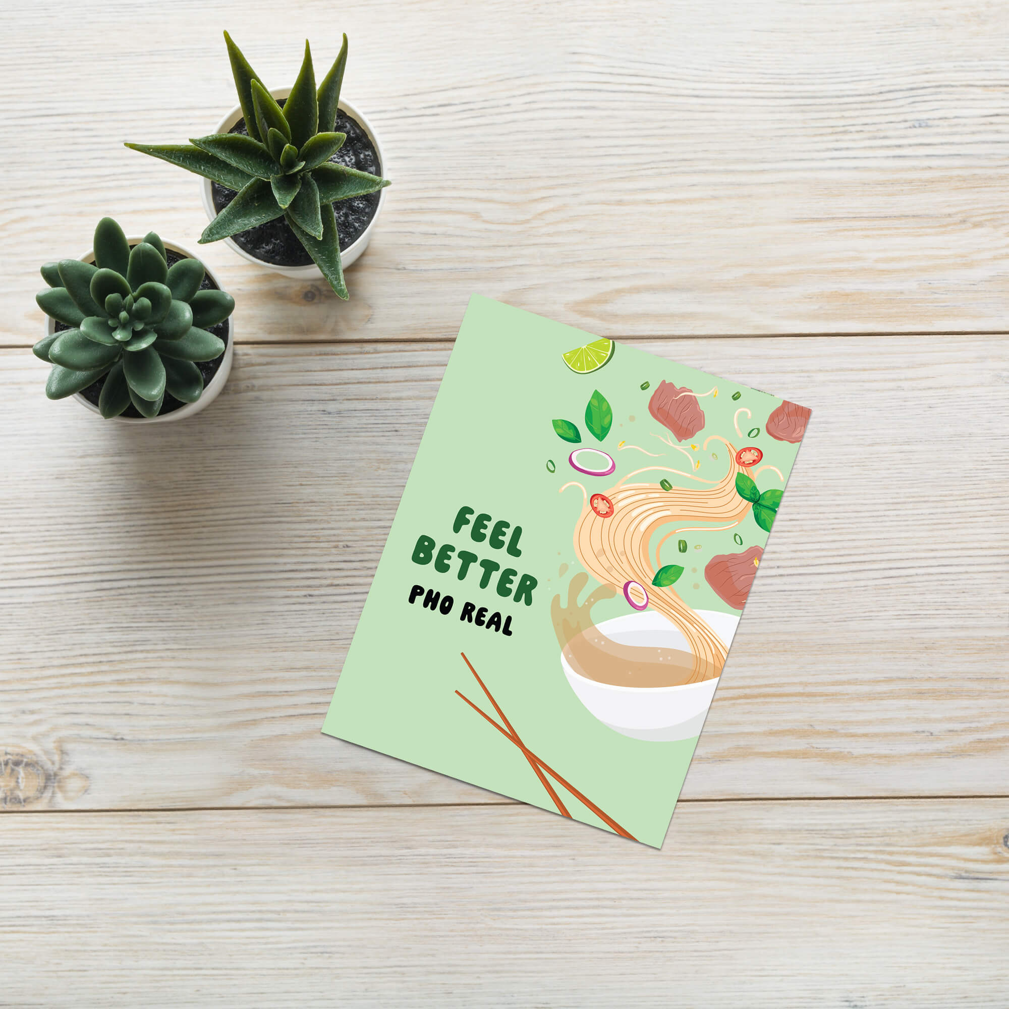 Greeting card with 'Feel Better Pho Real' text on a wooden surface with potted plants.