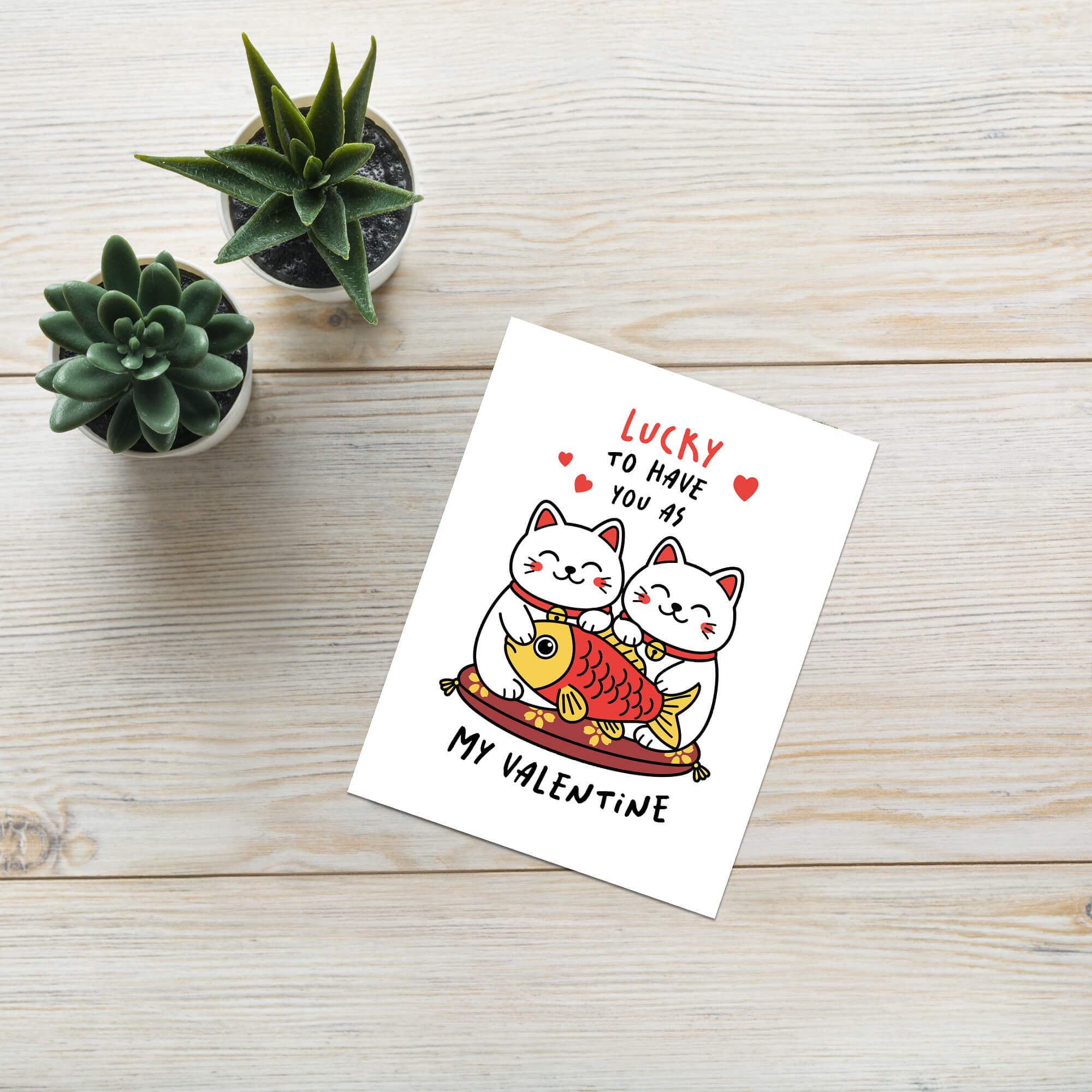 Valentine's Day card with two cats and fish on a wooden surface with small plants.