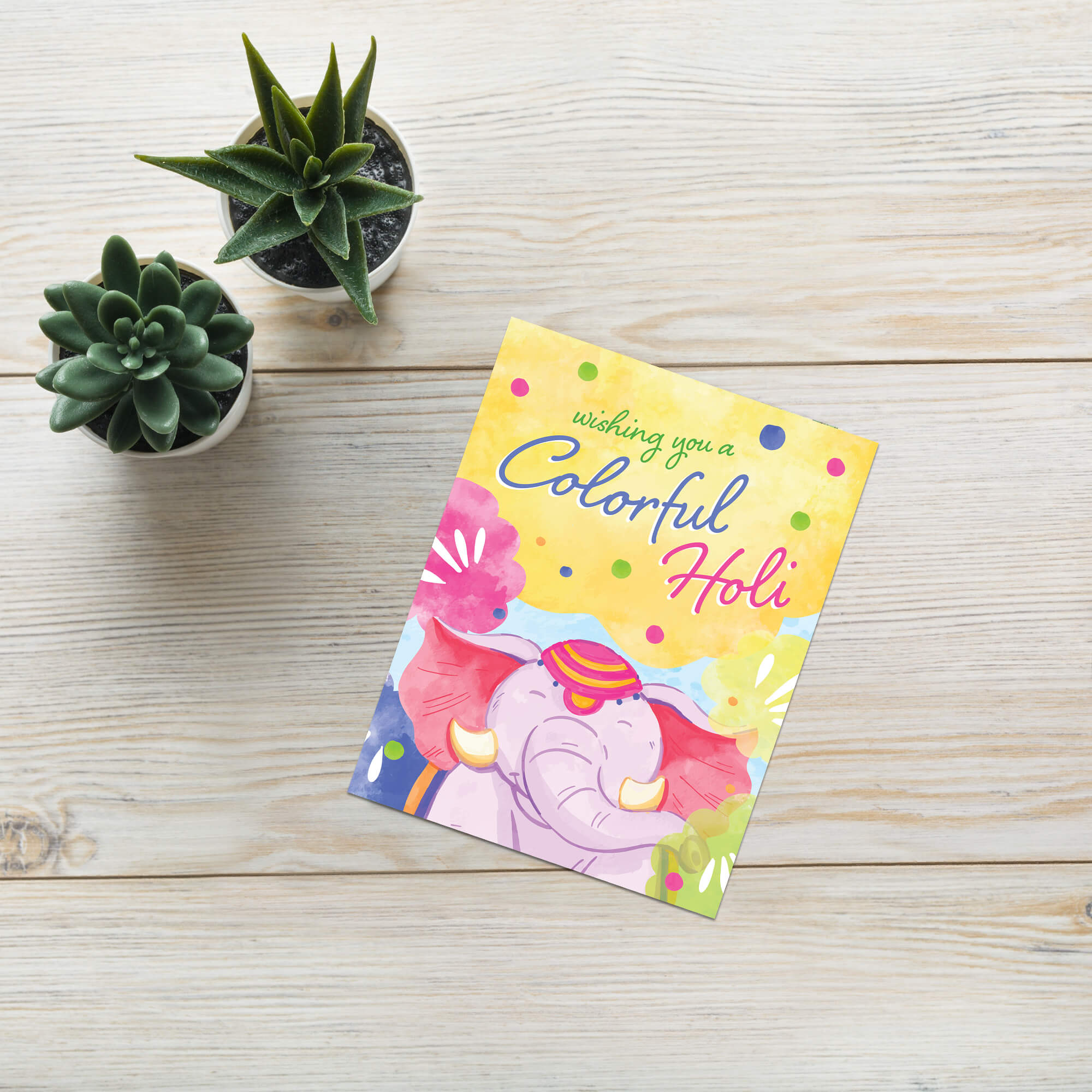 Colorful Holi greeting card with elephant design on a wooden surface with small potted plants.