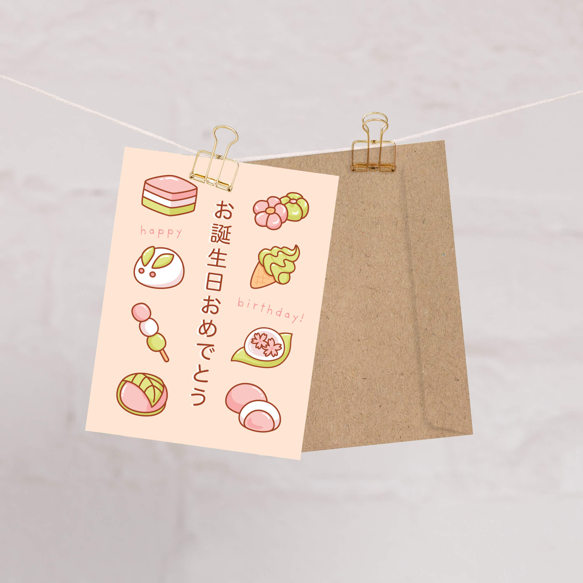 A Japanese birthday greeting card with cute illustrated sweets and the text 'happy birthday!' and 'ないじさすそじこじさす' on a pale pink background, clipped to a string with a kraft paper envelope behind it on a light marble-like surface.