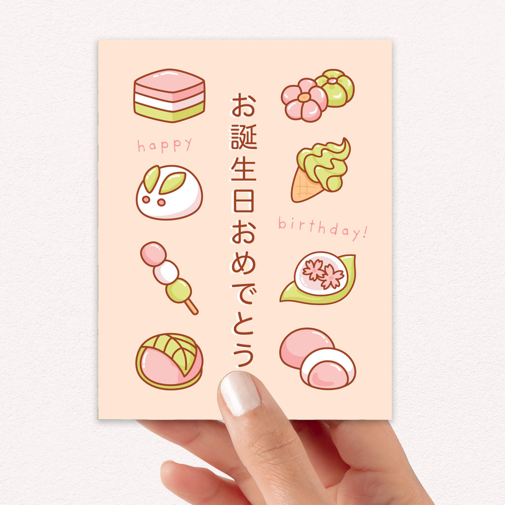 A hand holding a pastel birthday card featuring cute Japanese sweets and the text 'happy birthday!'. The card has a soft peach background with illustrations of pink, green, and white desserts, and is photographed against a light neutral wall.