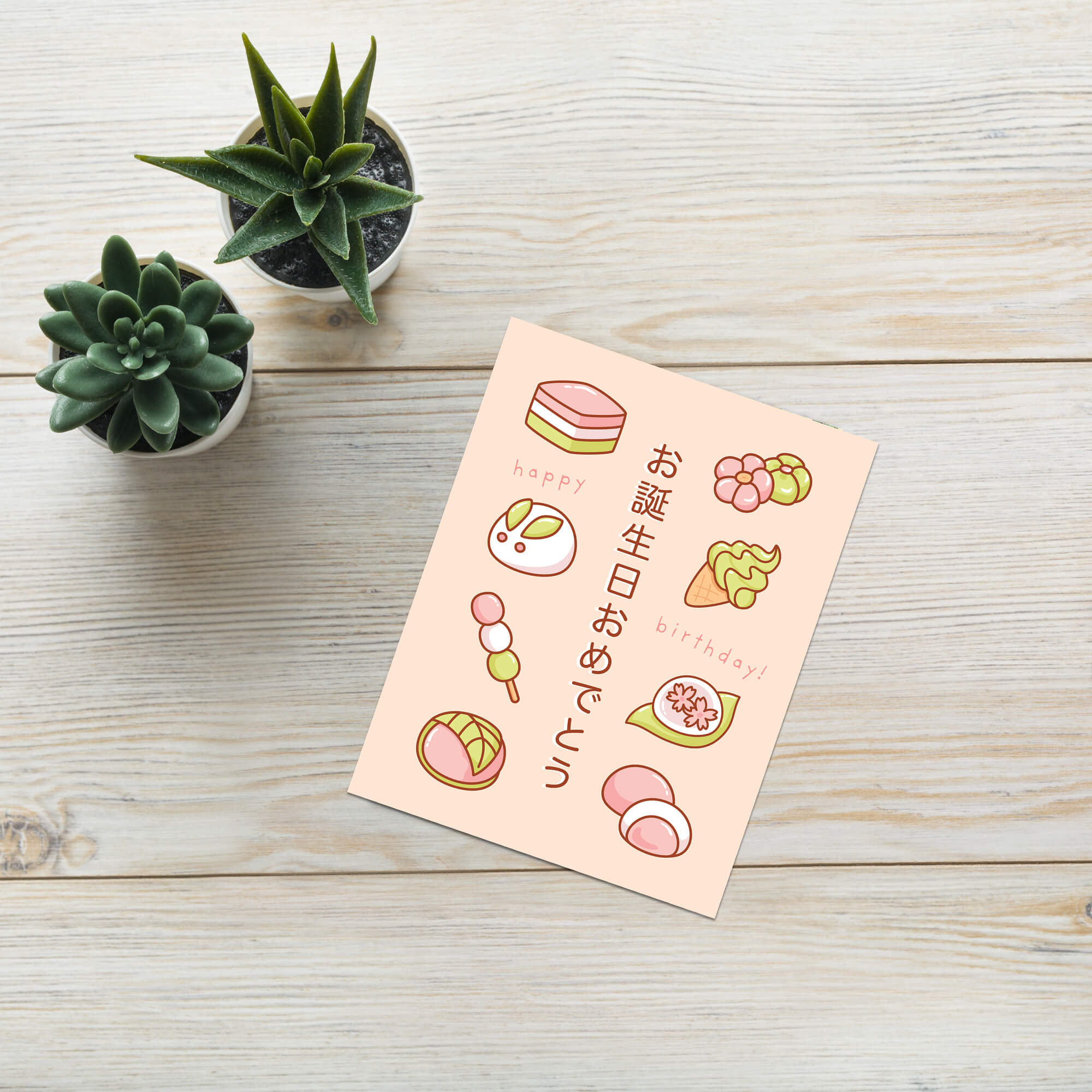 A Japanese birthday greeting card with cute illustrations of sweets and Japanese text, displayed on a light wood tabletop next to two small potted succulents.
