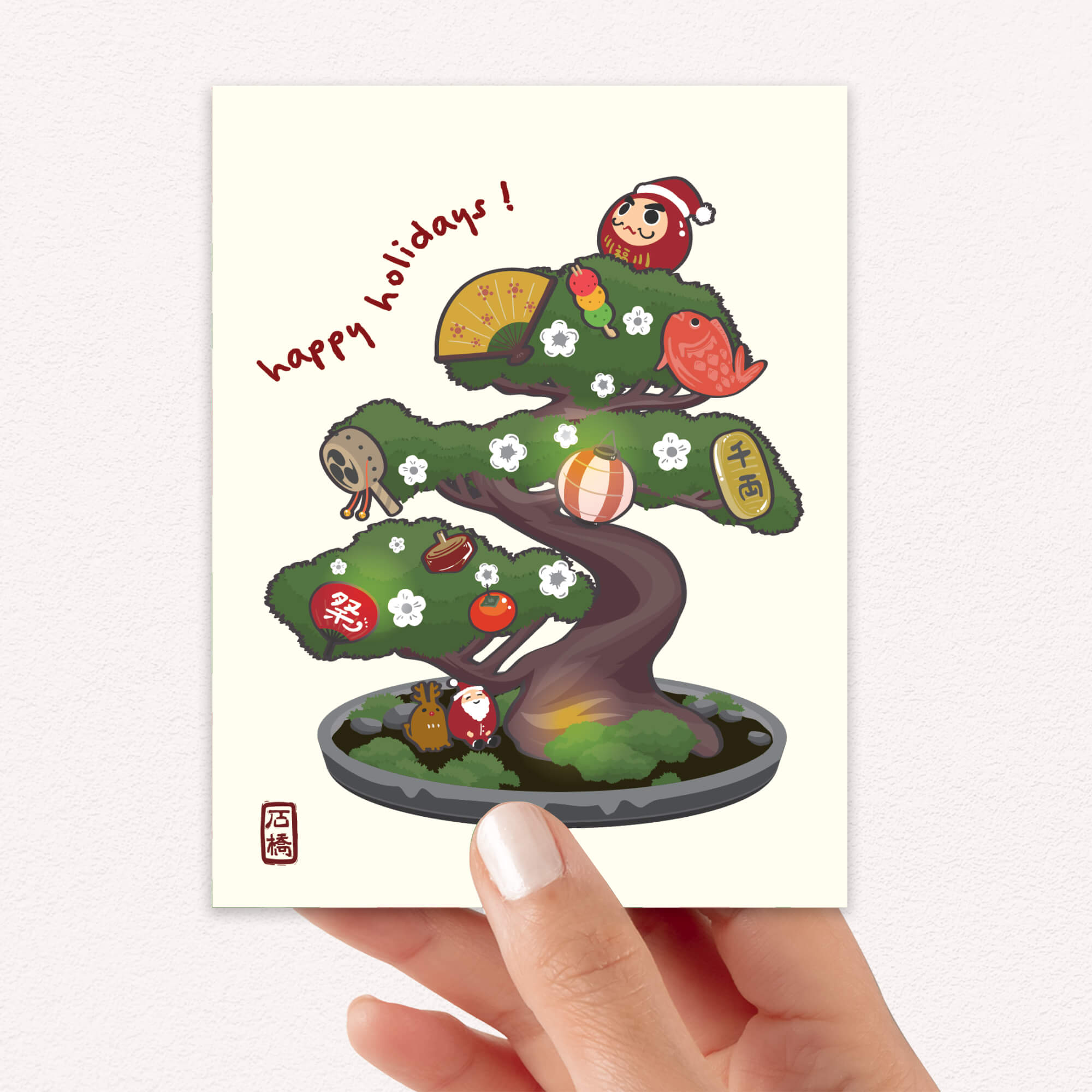 cute japanese christmas card with an Illustration of a bonsai tree decorated with lucky charms, daruma, and red lanterns. Text reads “happy holidays!”