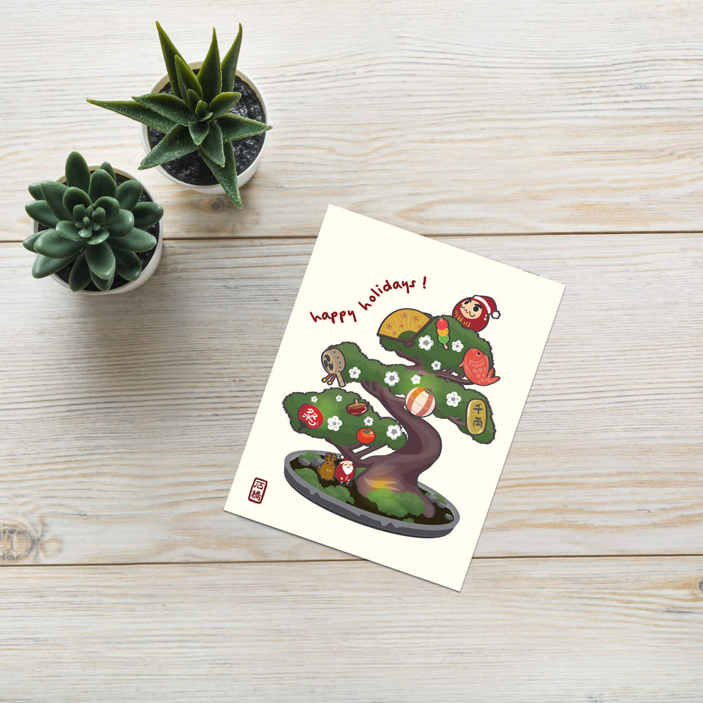 cute japanese christmas card with an Illustration of a bonsai tree decorated with lucky charms, daruma, and red lanterns. Text reads “happy holidays!”
