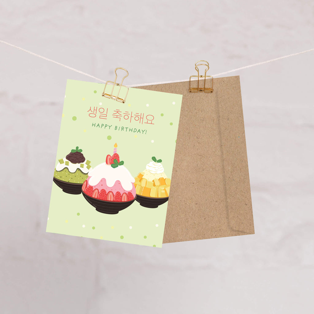 A Korean birthday card clipped to a string with a kraft paper envelope behind it. The card has a pale green background with three illustrated desserts and Korean and English text reading 'HAPPY BIRTHDAY'.