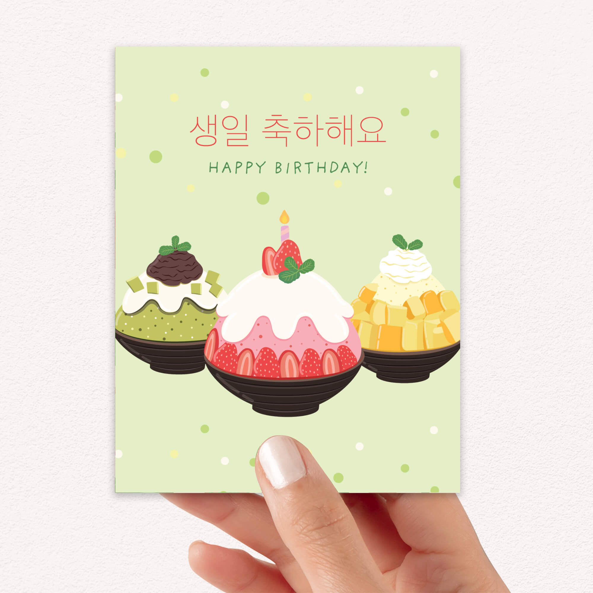 A hand holding a Korean birthday card with a light green background, Korean 'Happy Birthday' text, and three illustrated dessert illustrations in the center.