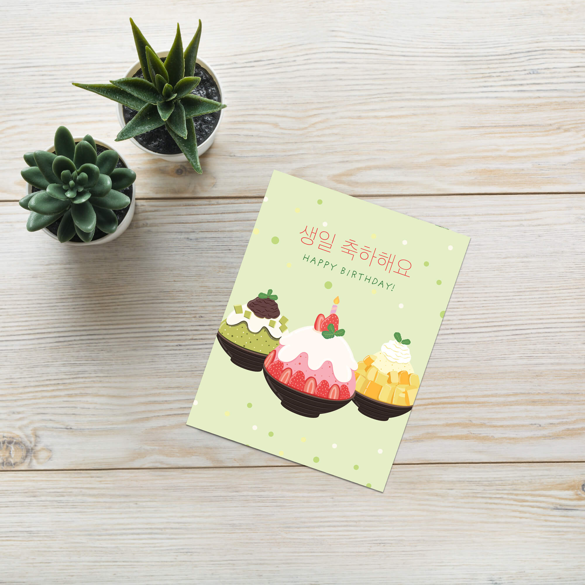 A light green birthday card with illustrations of three desserts and Korean text reading '생일 축하해요' and 'HAPPY BIRTHDAY!' lies on a light wood surface next to two small potted succulents.