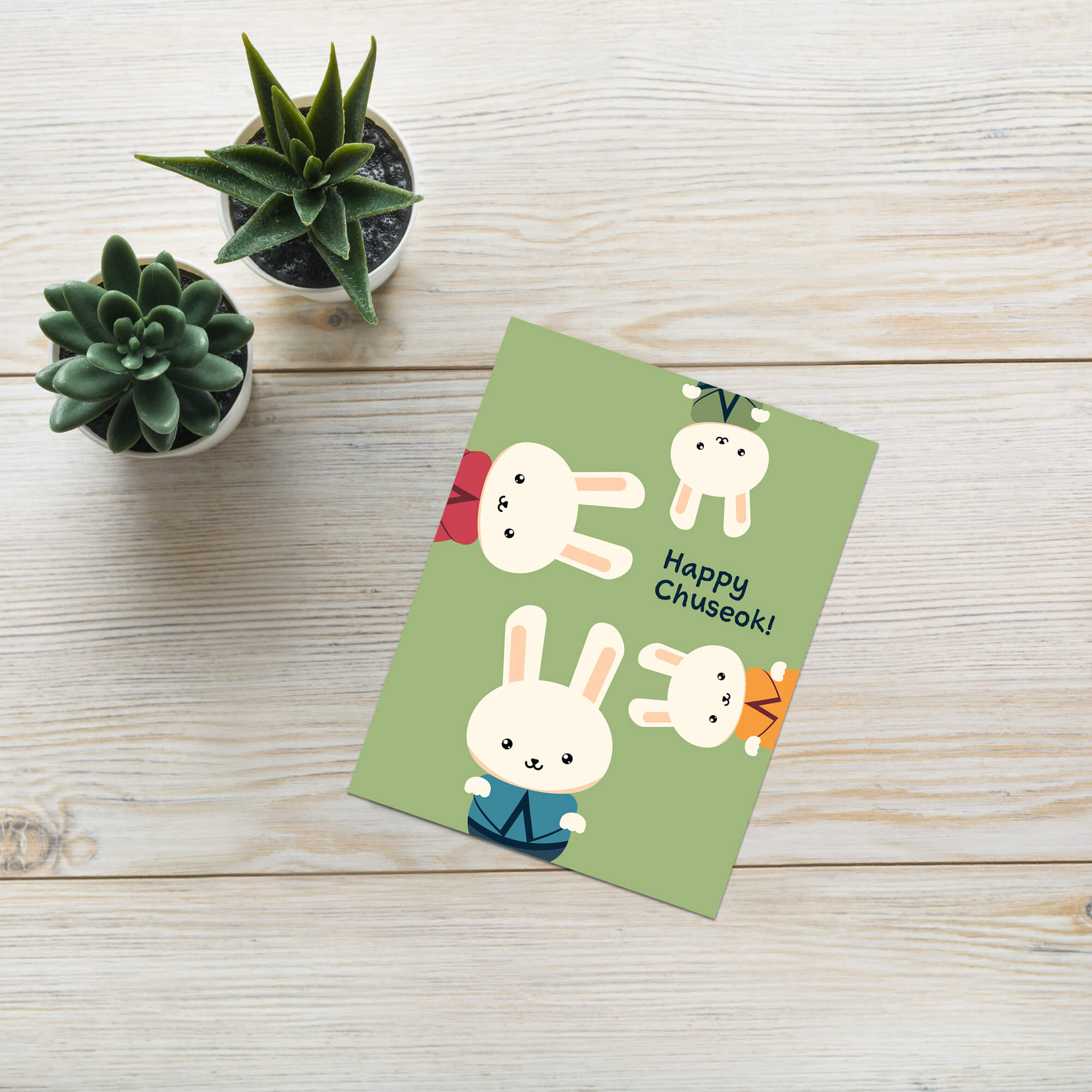 korean harvest thanksgiving greeting Green card with cartoon rabbits and 'Happy Chuseok!' text on a wooden surface with potted plants.