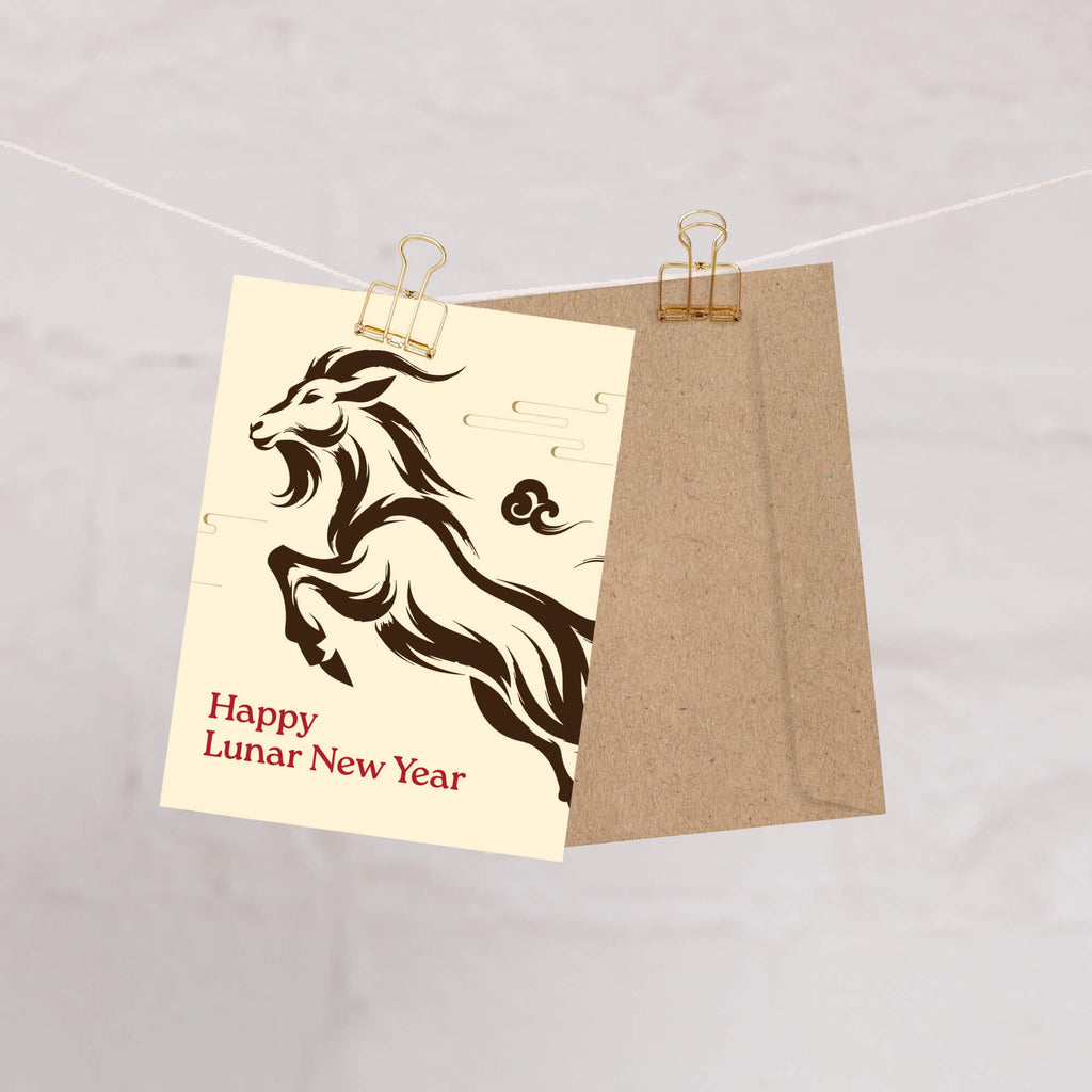 A Year of the Goat card with a black ram illustration and the text 'Happy Lunar New Year' in red, clipped to a string next to a brown kraft envelope against a light gray marble background.