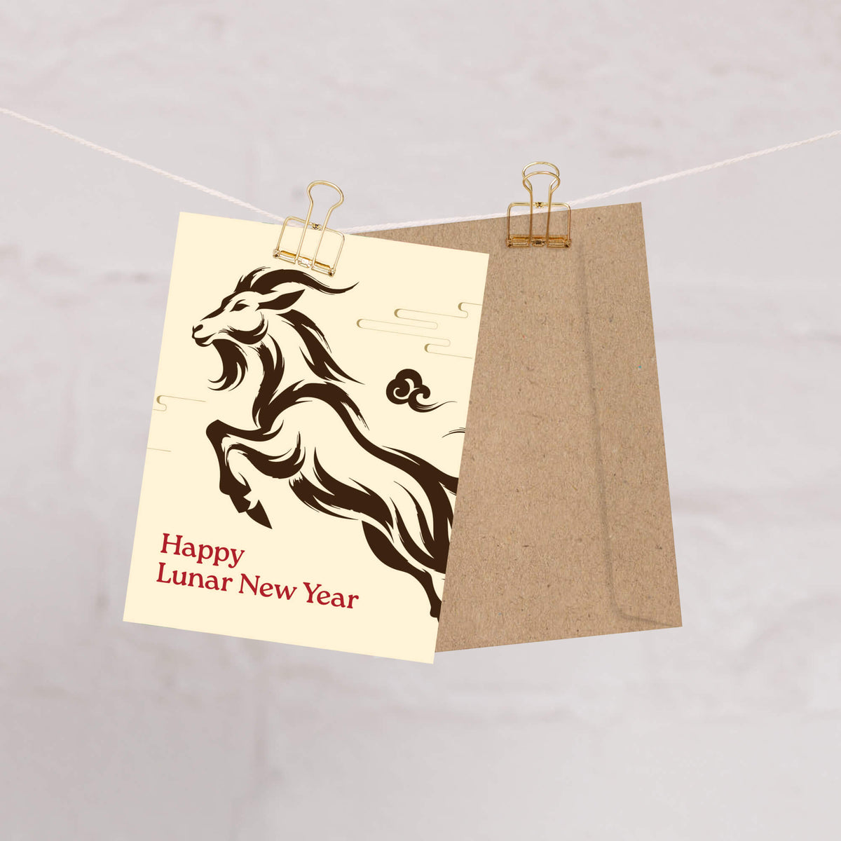A Year of the Goat card with a black ram illustration and the text 'Happy Lunar New Year' in red, clipped to a string next to a brown kraft envelope against a light gray marble background.