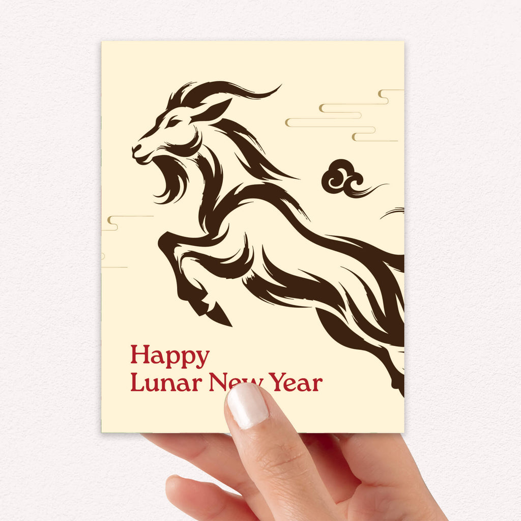 A hand holding a cream-colored chinese new year card featuring a dark brown stylized galloping goat and the text 'Happy Lunar New Year' in red, against a light textured background.