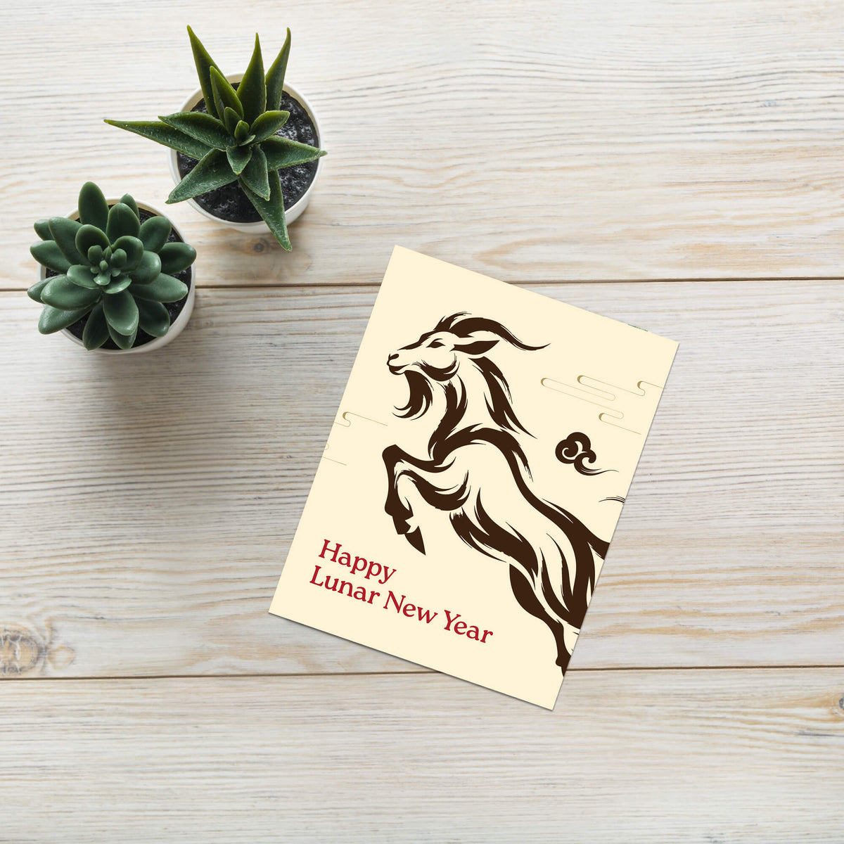 A square lunar new year card with a dark brown goat illustration and the text 'Happy Lunar New Year' in red, placed on a light wood surface next to two small potted succulents.